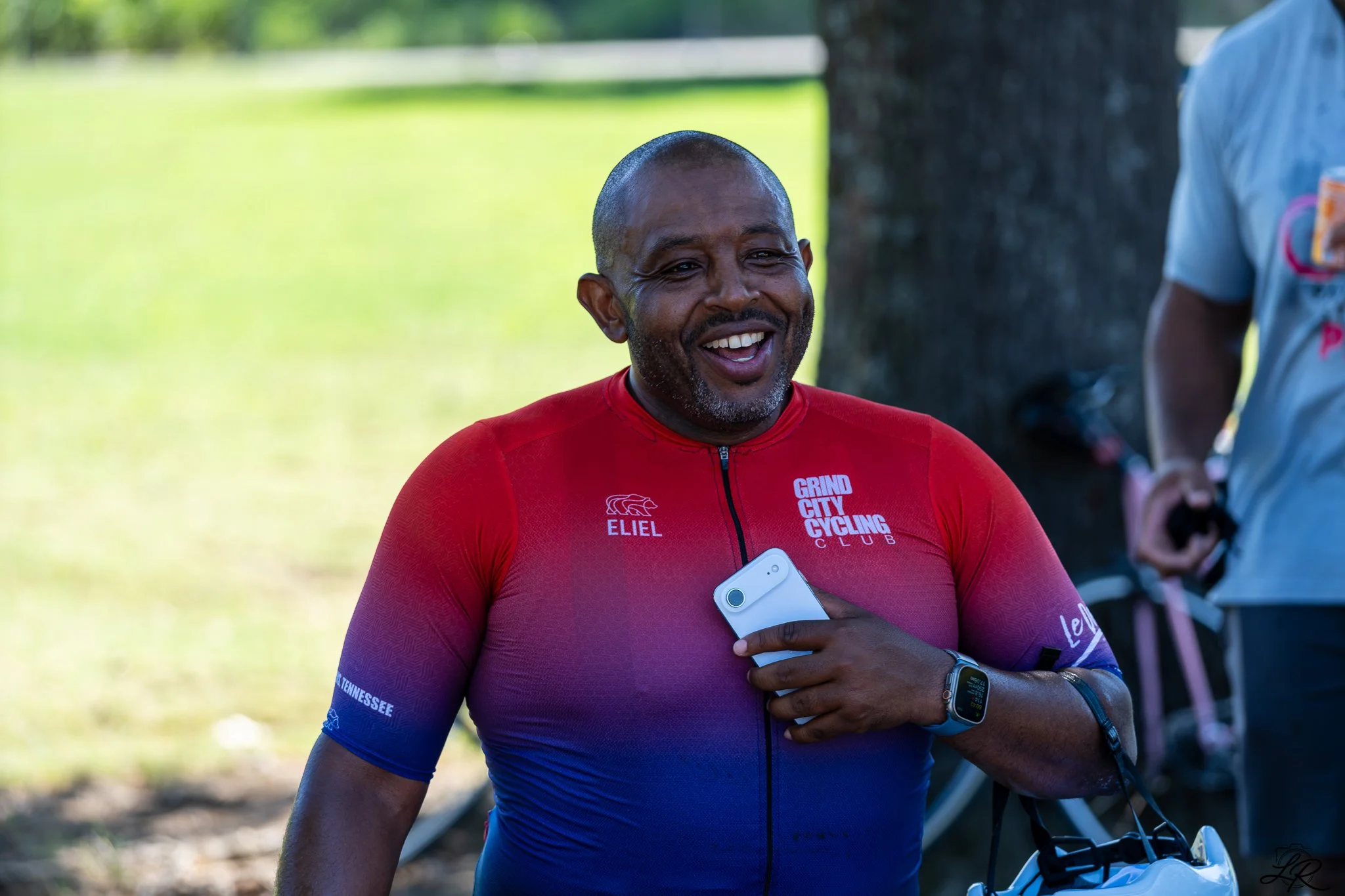 A man smiling outdoors wearing a red and blue cycling jersey holding a smartphone, standing next to a bicycle, with a tree and grassy area in the background.