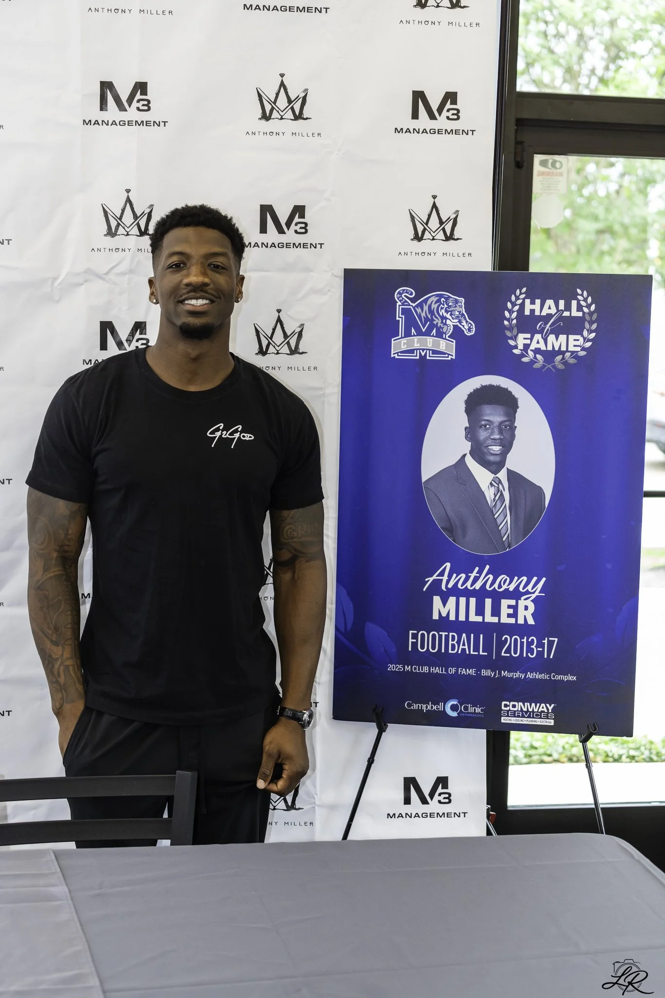 Young man standing in front of a backdrop with logos, wearing a black t-shirt and smiling. To the right, there is a blue poster with a black and white photo of him, indicating he played football from 2013 to 2017, and featuring the Hall of Fame logo.