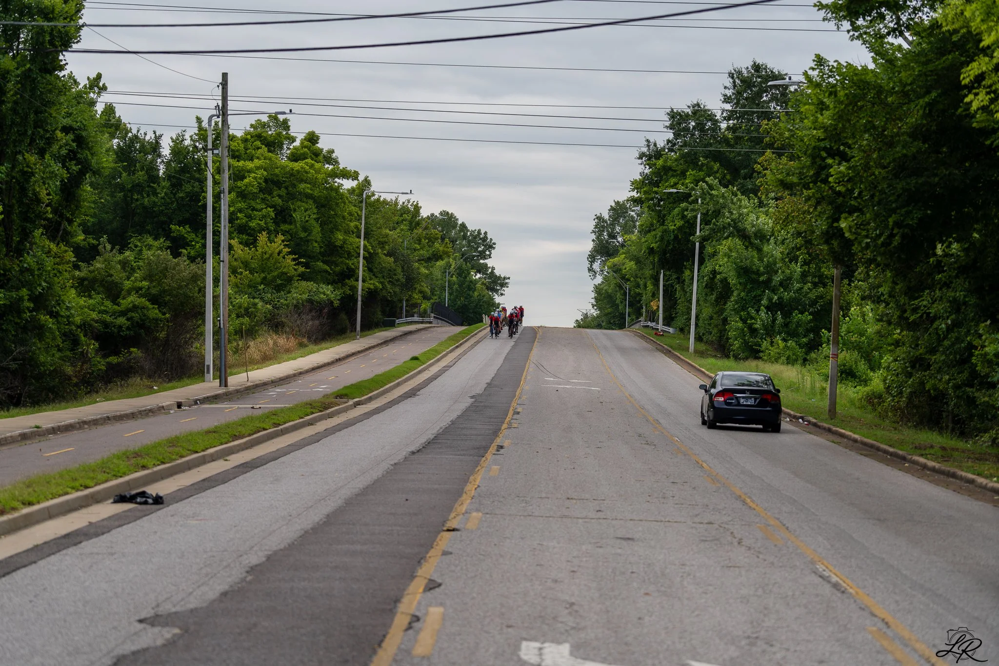 A straight road with a bicycle lane on the left side, surrounded by lush green trees and overcast sky. A group of cyclists is riding in the distance, and a black car is parked on the right side of the street.