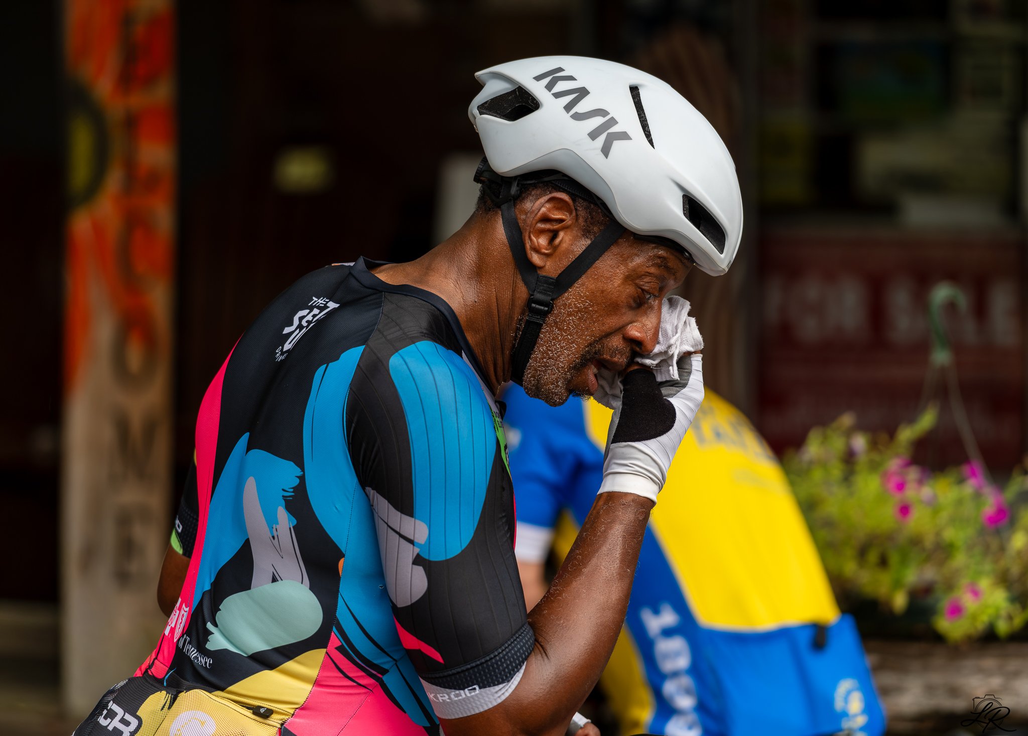 An older man wearing a white bike helmet and colorful cycling jersey, wiping sweat from his face with a cloth, in a setting with blurred banners and flowers in the background.