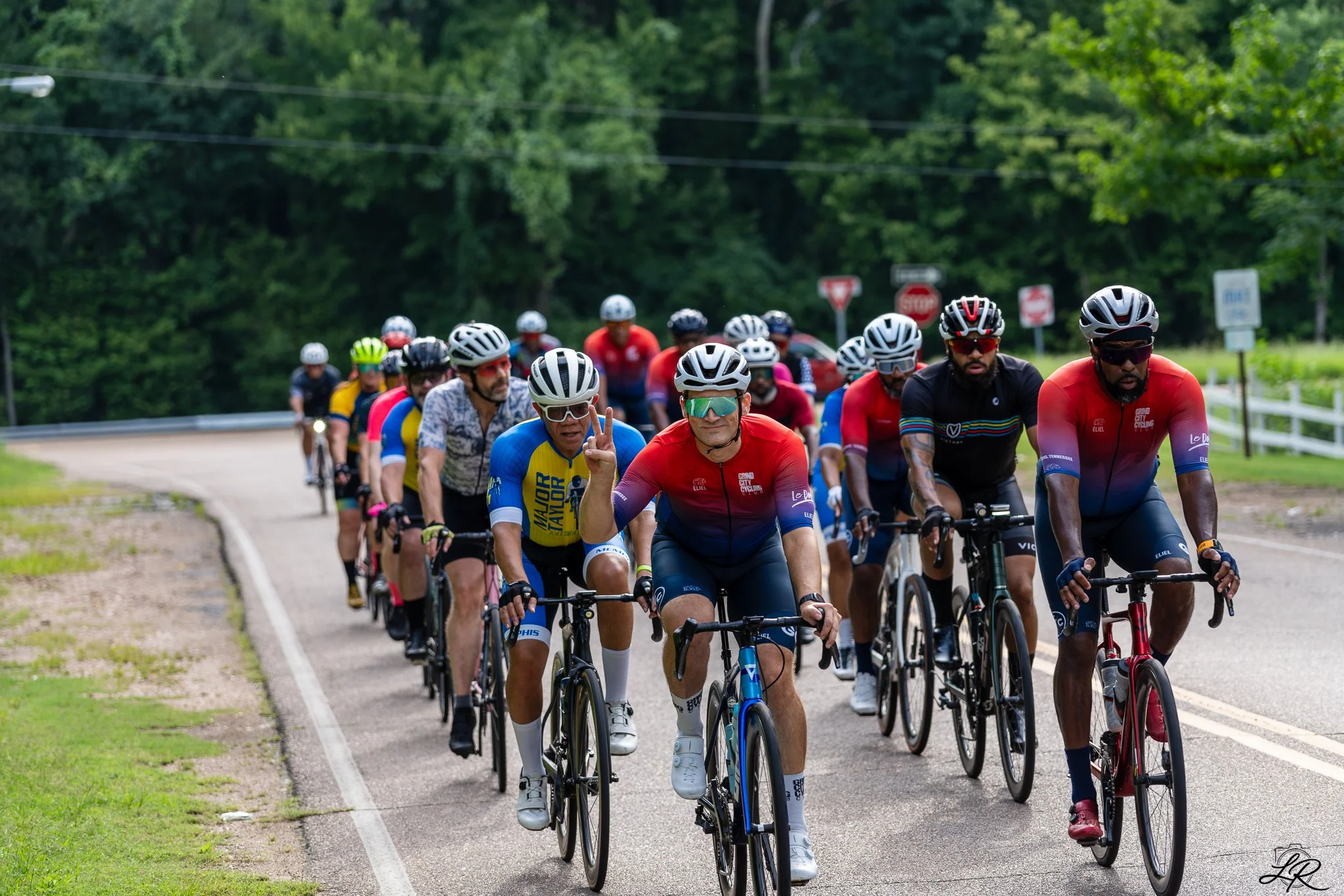Group of cyclists riding on a paved road surrounded by green trees, some making peace signs, wearing athletic gear and helmets.