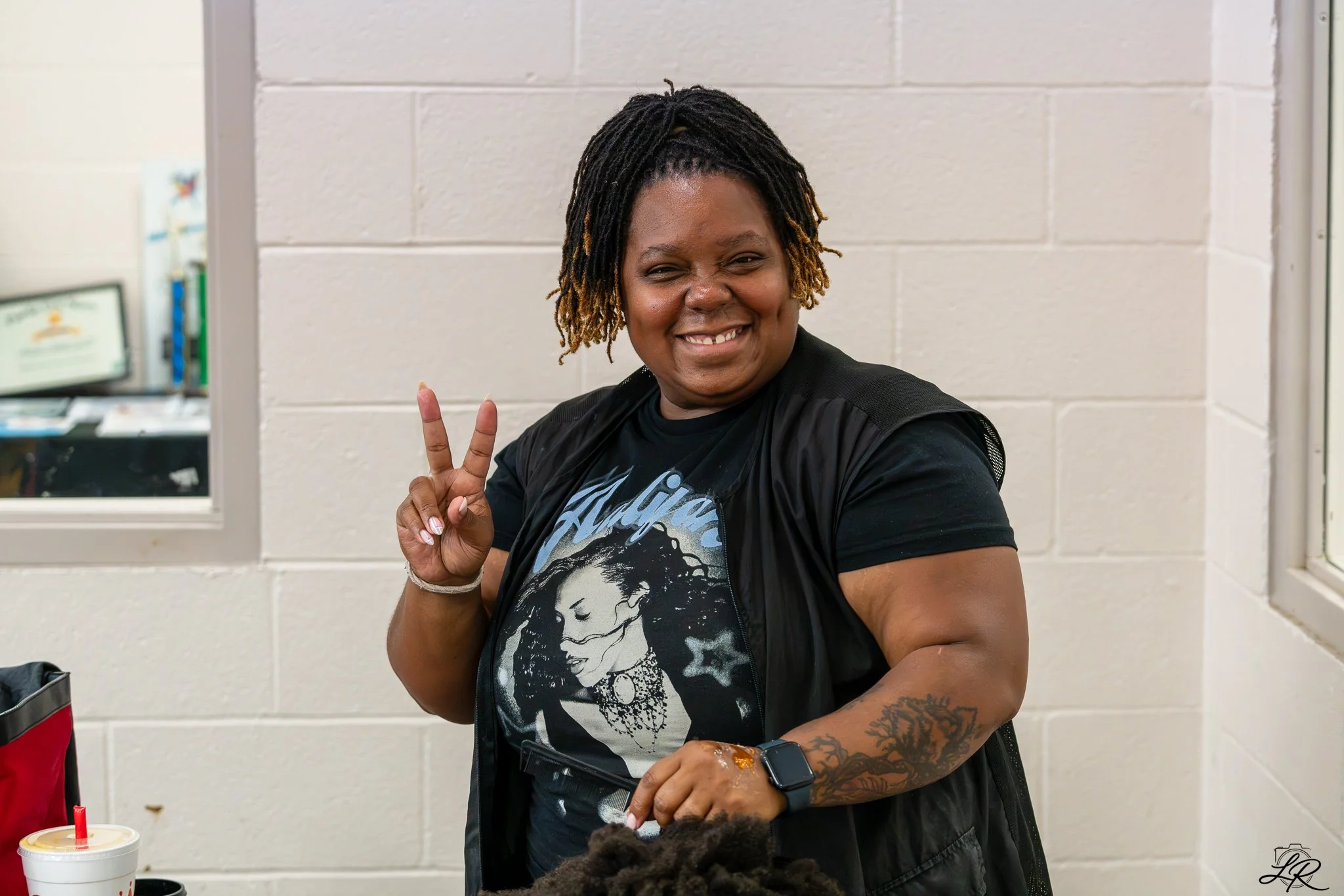 A woman with dark skin and dreadlocks making a peace sign with her hand smiling at the camera. She is at a table with a drink and has tattoos on her left arm. The background has a white brick wall and a window.
