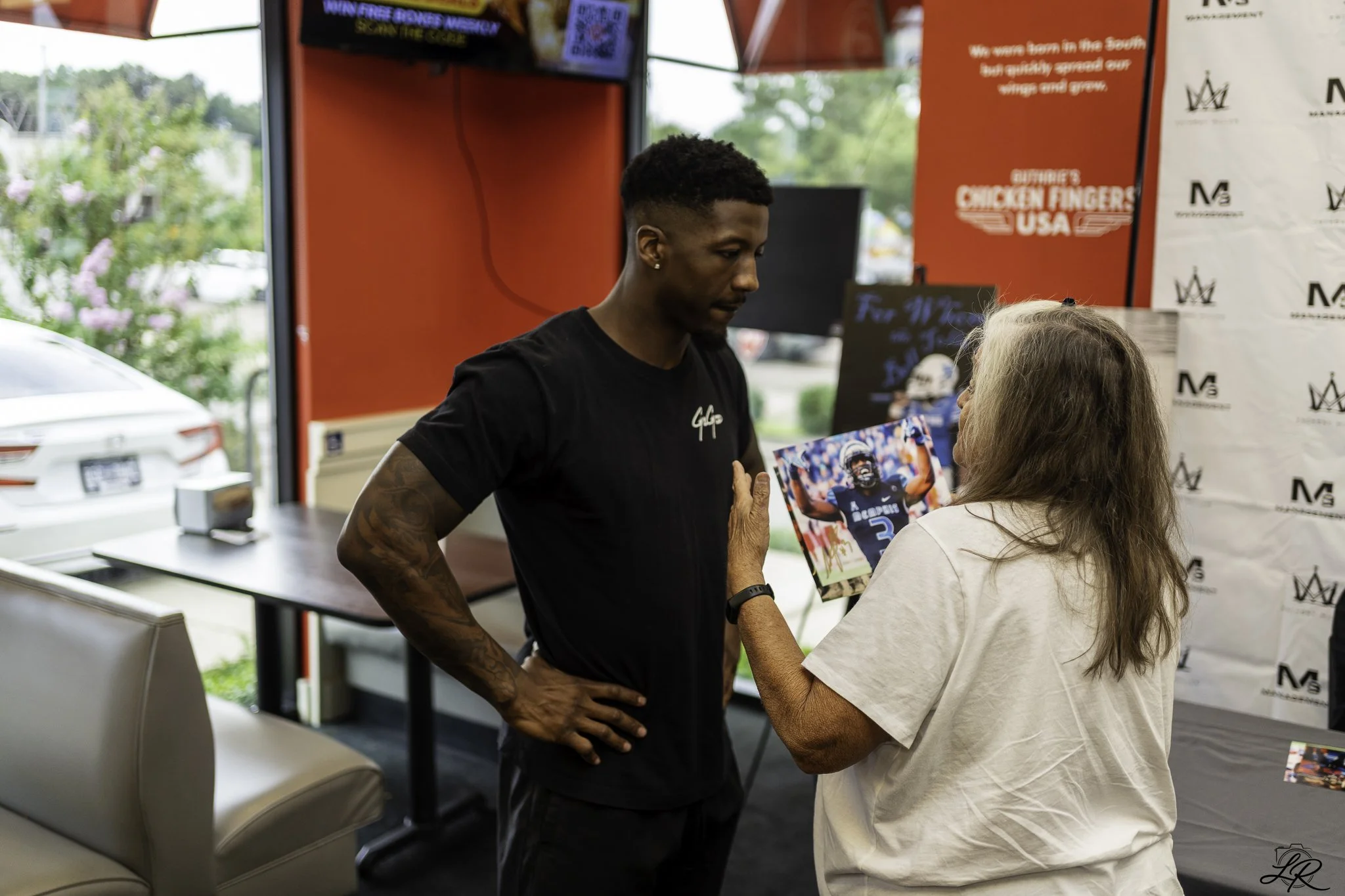 A person with dark skin, short hair, wearing a black shirt, is talking to a woman with long light hair, outside of a stall with red and white walls. The person is holding a colorful picture of a football player.