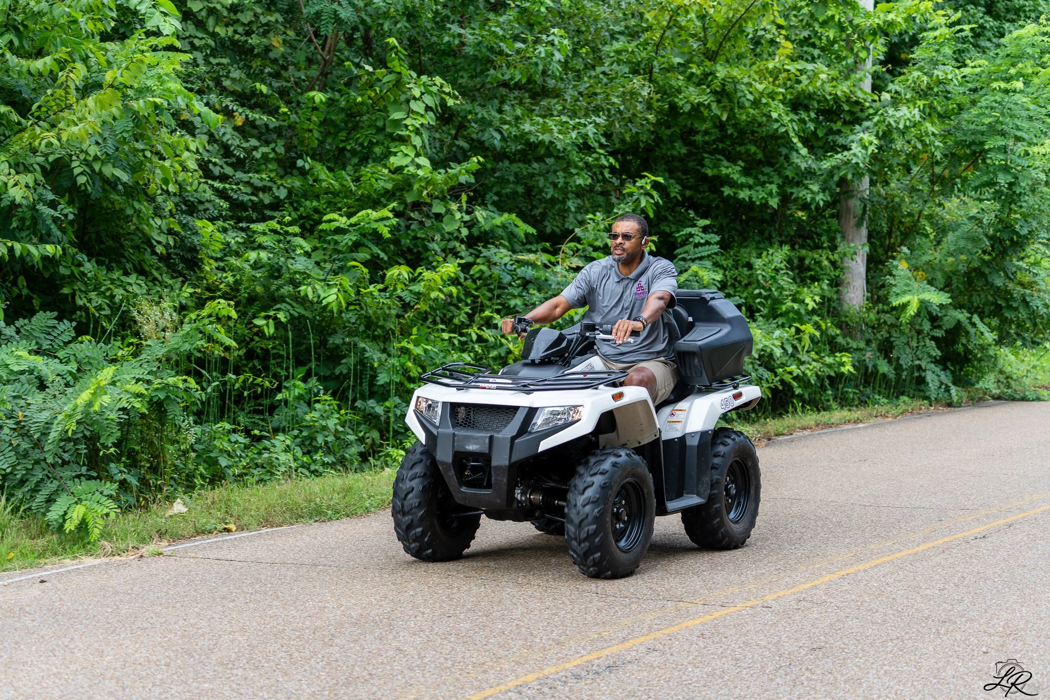 A man riding an all-terrain vehicle (ATV) on a paved road beside green trees and bushes. The man is wearing sunglasses, a gray polo shirt, and khaki shorts.
