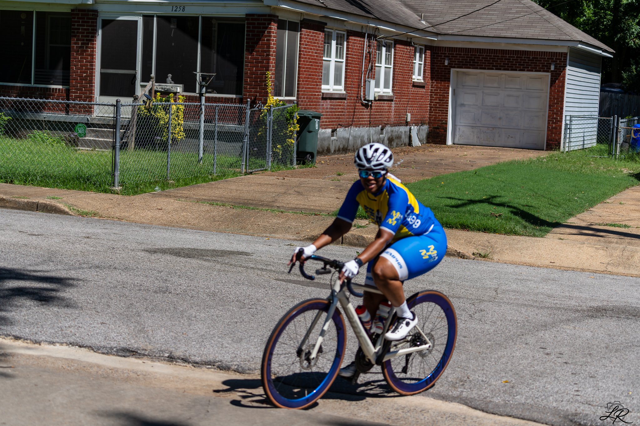 A young boy wearing a yellow and blue cycling jersey, a white helmet, and sunglasses riding a white mountain bike on a suburban street in front of a brick house with a garage. The street has a curb, and the house has a chain-link fence, a small grass