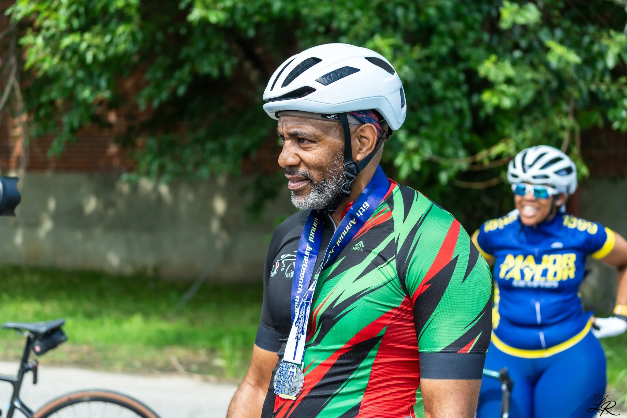 A man wearing a black and green cycling jersey, a white bike helmet, and a medal around his neck, standing outdoors with a woman in a blue cycling jersey and helmet smiling in the background.