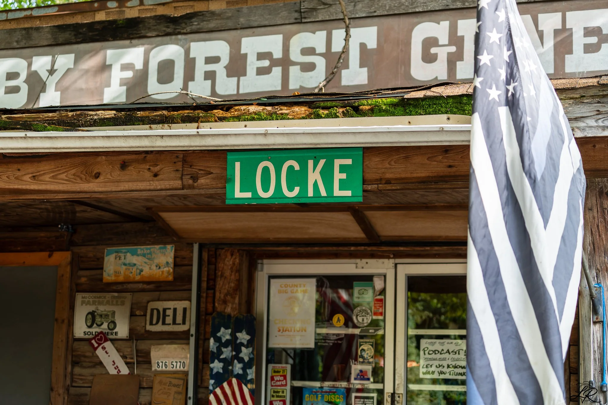 A rustic wooden storefront with a green 'LOCKE' sign, posters, and a partial American flag hanging. The shop appears to sell various items including golf discs, and has nostalgic signs like one for a tractor and deli, under a sign that says 'BY FORES