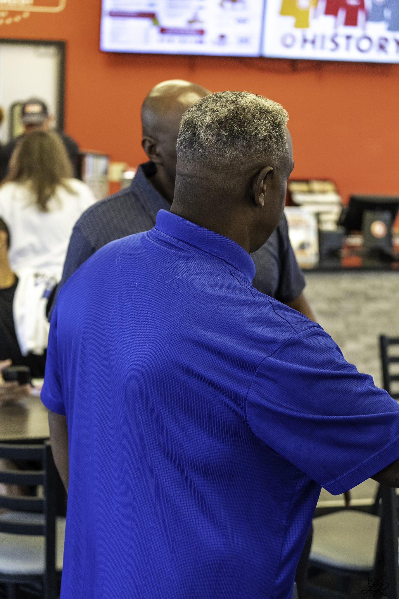 A man with curly gray hair wearing a bright blue shirt, viewed from behind, standing in a busy indoor setting with red walls, people, and computer monitors in the background.