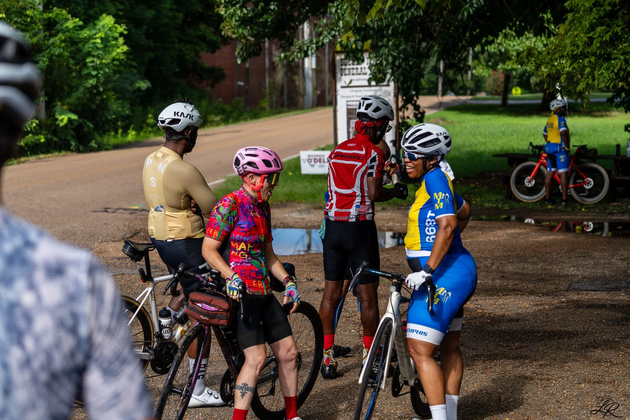 Group of cyclists wearing helmets and colorful attire gathered in a park, smiling and chatting, with trees and a road in the background.