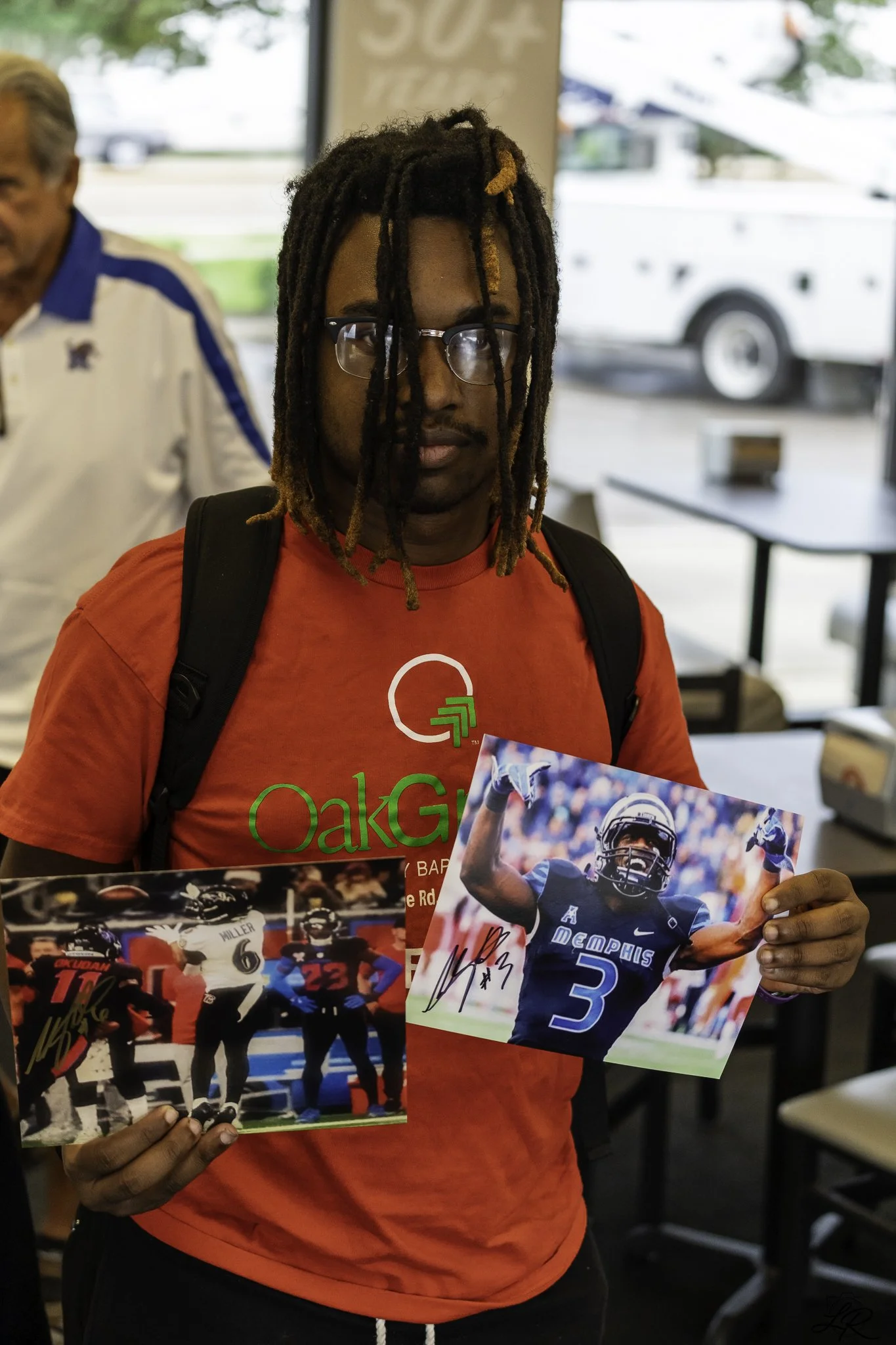 A man with dreadlocks wearing glasses and a red shirt holding signed sports photos, likely of football players, in a casual indoor setting.