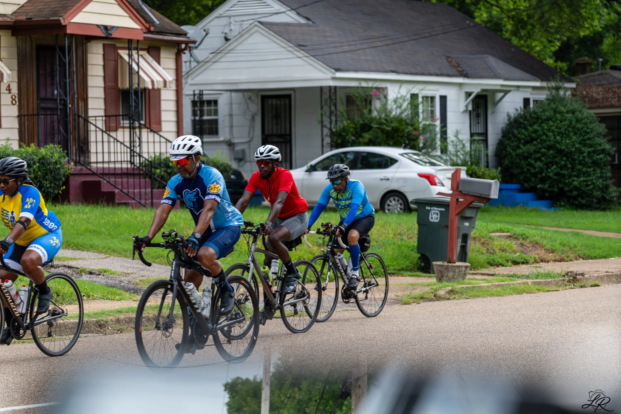 A group of people in cycling gear riding bikes on a residential street with houses and cars in the background.
