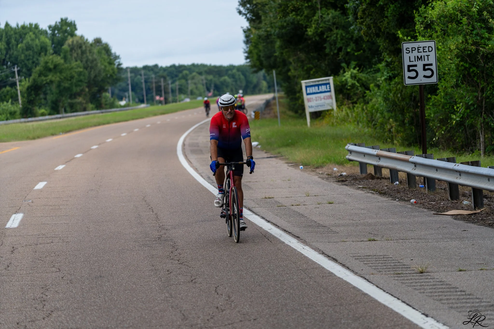 Man in red cycling jersey and helmet riding a road bike along a rural highway with a 55 mph speed limit sign on the side of the road and trees in the background.