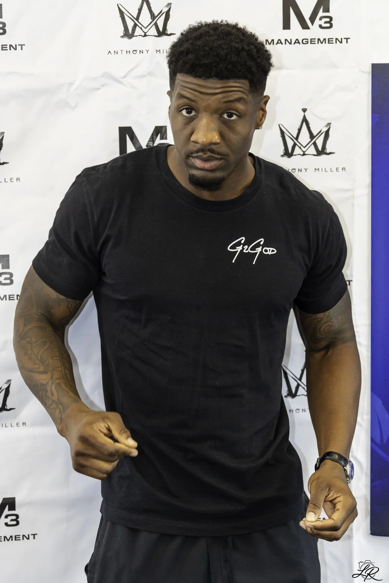A young man with short curly hair and tattoos on his arm, wearing a black GIGOD t-shirt and a wristwatch, making a fist with one hand and pointing with the other, standing in front of a white backdrop with logos including Anthony Miller, M3 Managemen