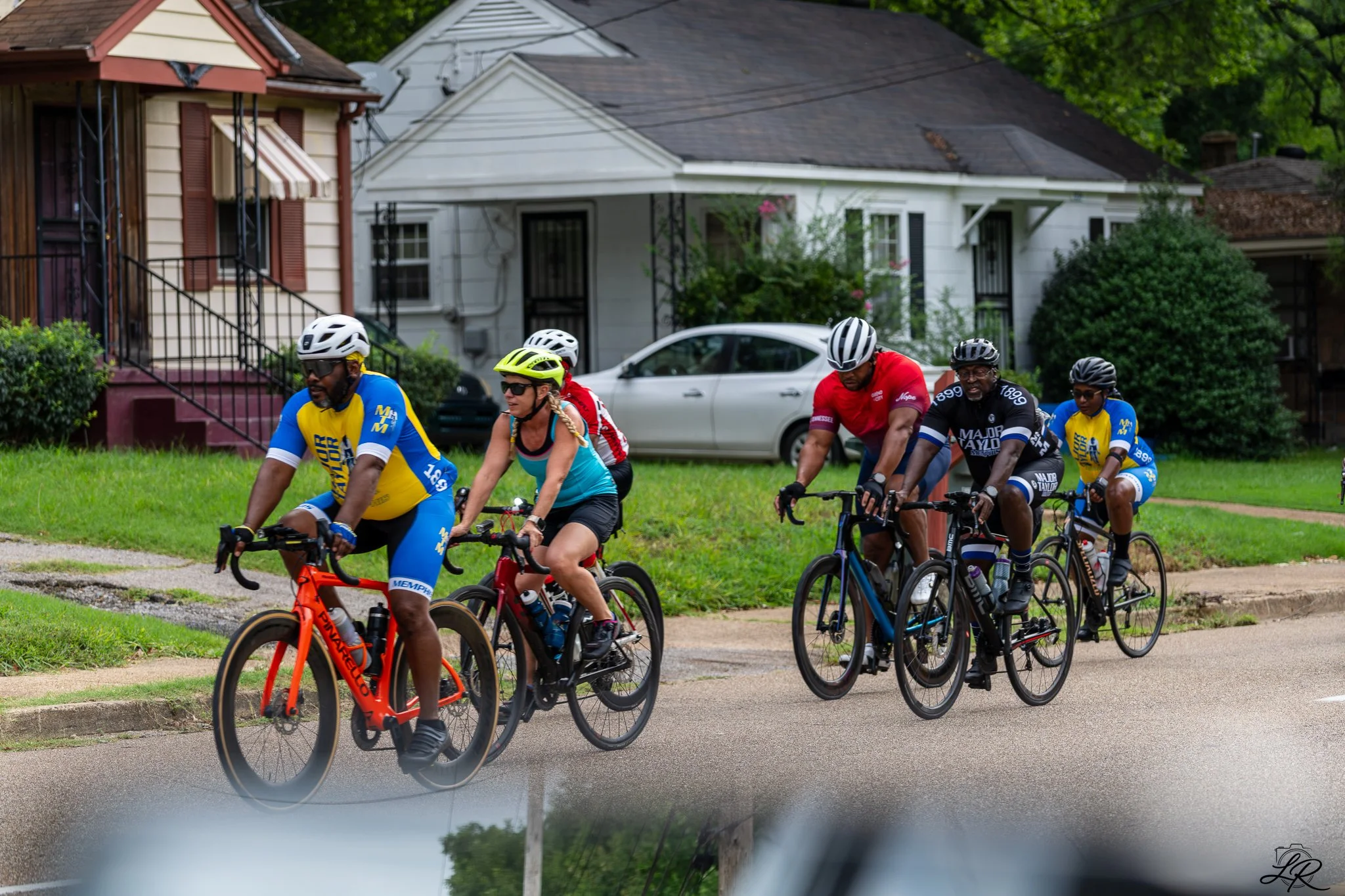 Group of cyclists riding on a residential street in front of houses and parked cars.