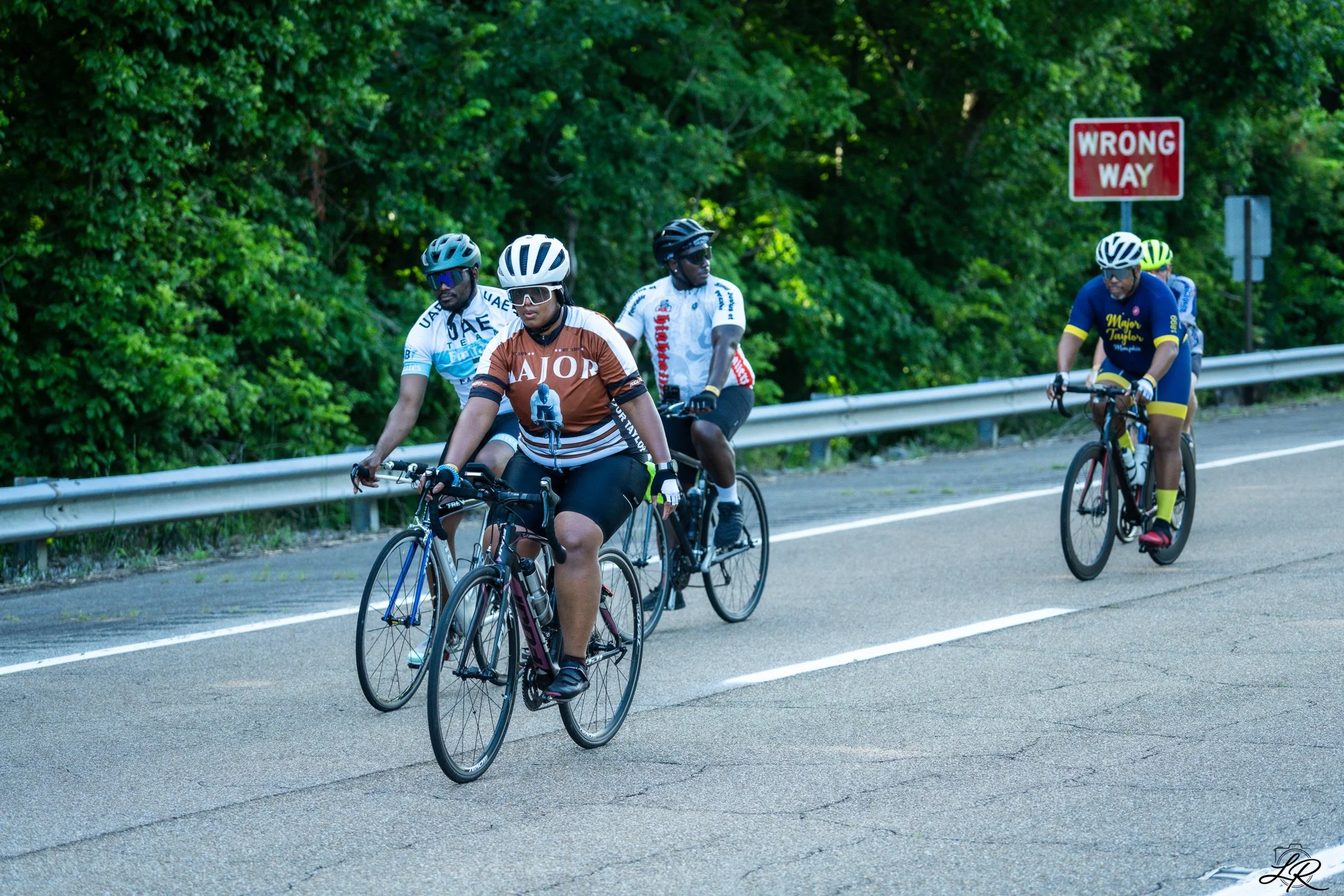 A group of five cyclists riding on a paved bike path beside a guardrail and dense green trees, with a red 'Wrong Way' road sign in the background.
