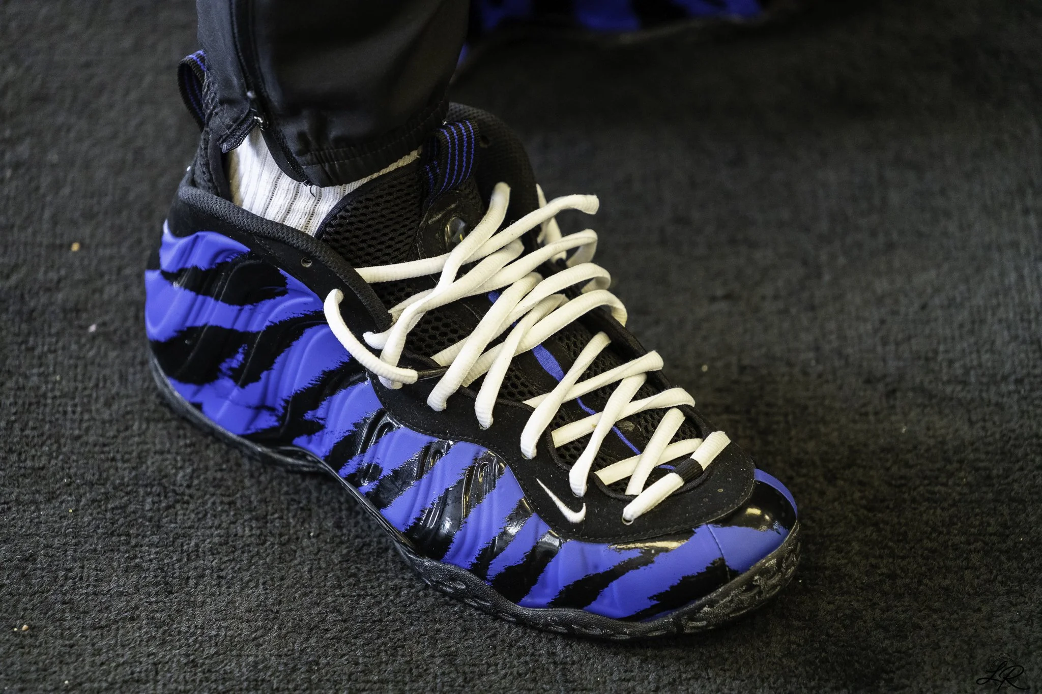 Close-up of a black and blue athletic shoe with white laces, worn with gray socks and black pants, on a dark textured floor.