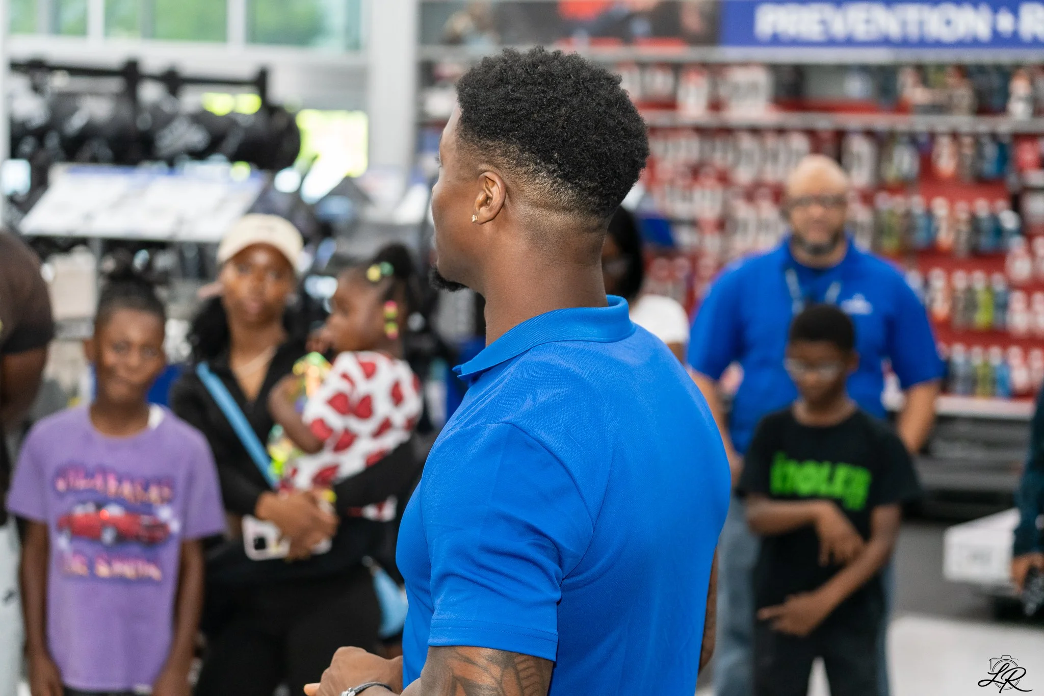 A man with short curly hair and earrings wearing a blue shirt is standing with his back to the camera in a store, surrounded by children and adults, with shelves of various products in the background.