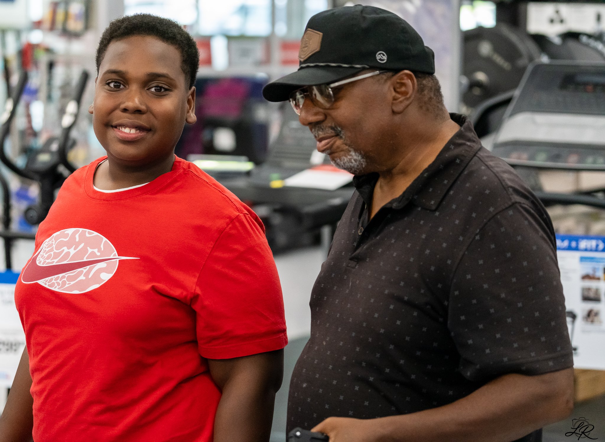A young boy and an older man, possibly grandfather, standing together in a store, smiling. The boy is wearing a red t-shirt with a graphic design, and the man is wearing a black cap and dark shirt with small patterns. They are near exercise equipment