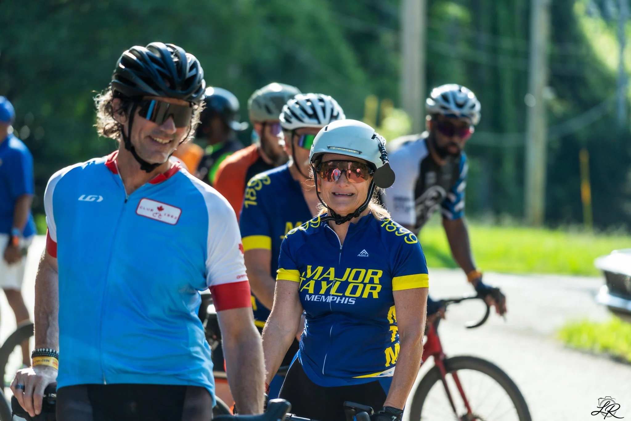 A group of people wearing cycling gear, helmets, and sunglasses, gathered outdoors on a sunny day with trees and power lines in the background. The woman in the center is wearing a blue and yellow jersey that says "Major Taylor Memphis," while the ma
