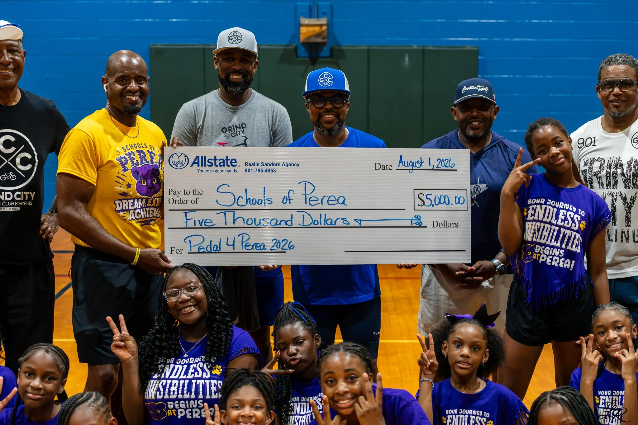 Group of smiling individuals, including children and adults, holding a large check for five thousand dollars made out to Schools of Perea, dated August 1, 2026, inside a gymnasium with blue walls.