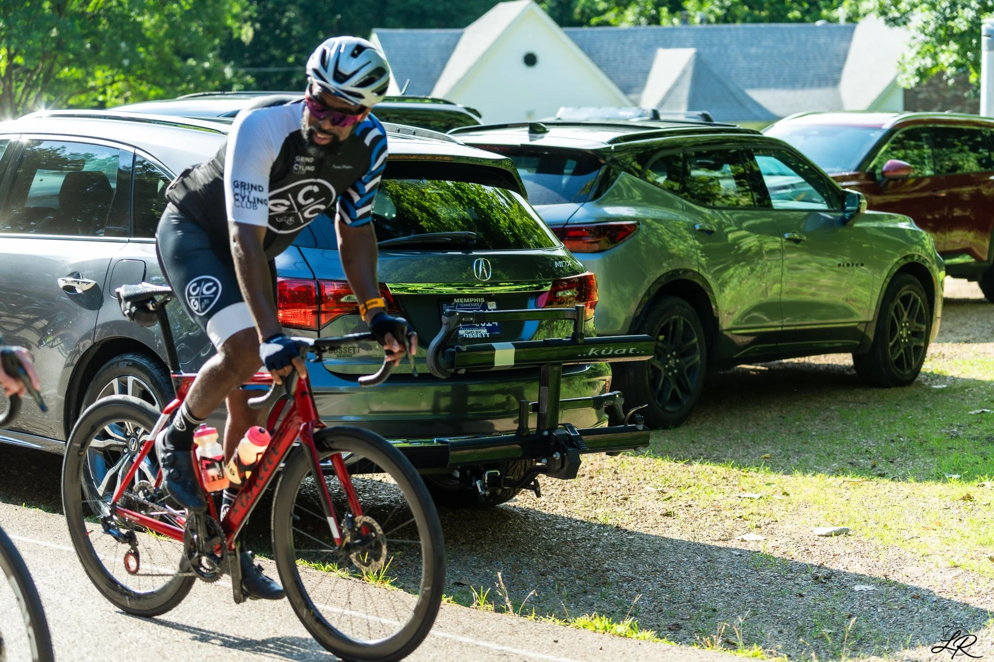 A cyclist wearing a helmet and sunglasses, riding a red bike with water bottles, passing parked cars, and trees in the background.