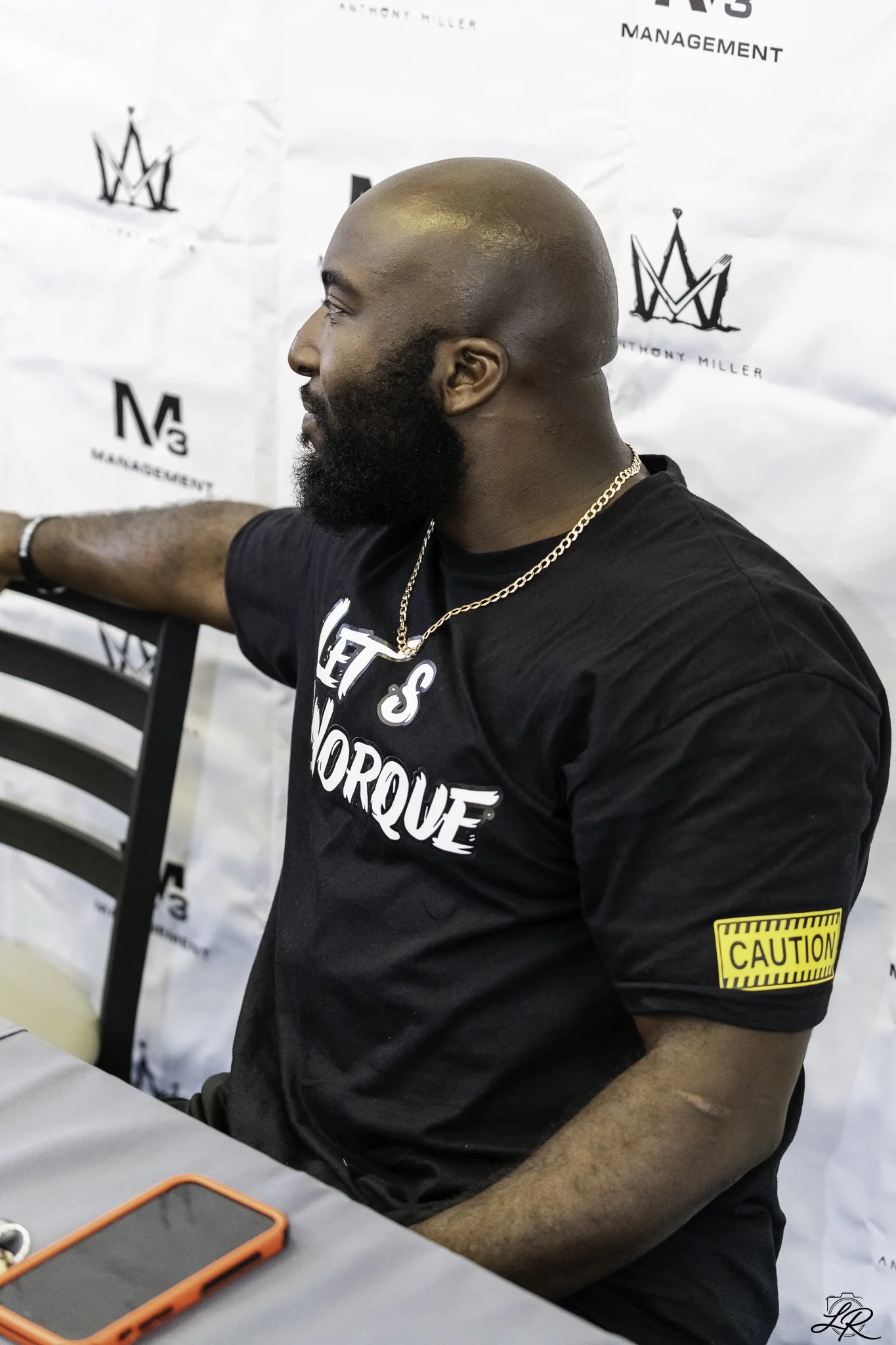 A man with a bald head and full beard, wearing a black T-shirt with white text, a gold chain, and a yellow caution armband, sitting at a table with a white background that has a black logo and text. There is a red phone case and earbuds on the table.