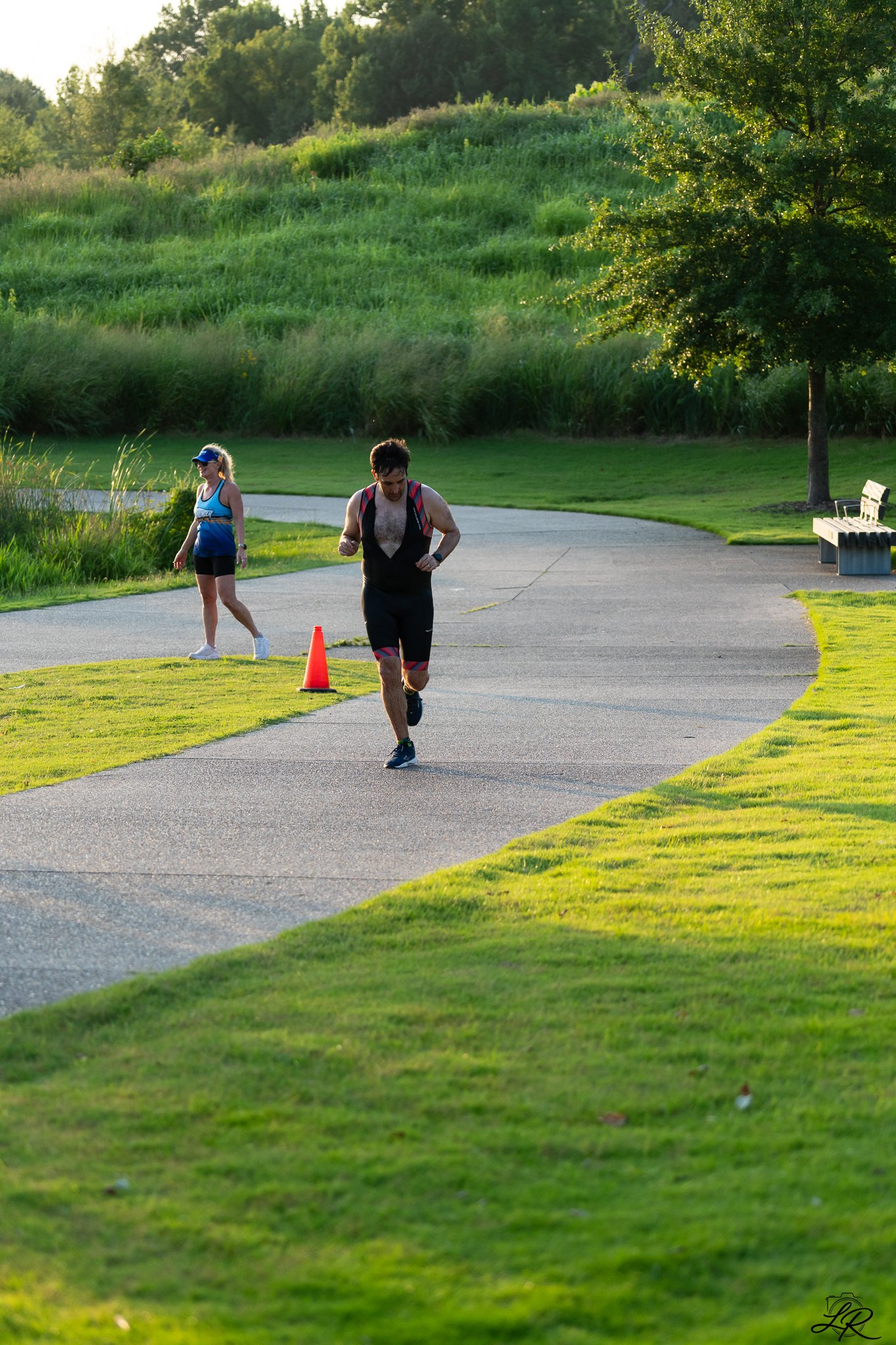 A man and a woman running outdoors on a paved path in a park during late afternoon. The man is shirtless, wearing shorts and a vest, while the woman is dressed in a tank top, shorts, and a cap. There is green grass and trees, with a large tree on the