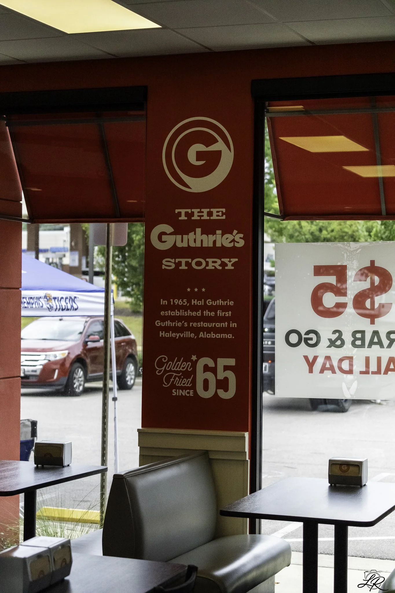 Interior view of a restaurant with tables and chairs, a red wall with a sign about The Guthrie's Story, a window showing parked cars outside, and a partial view of the restaurant's exterior signage.