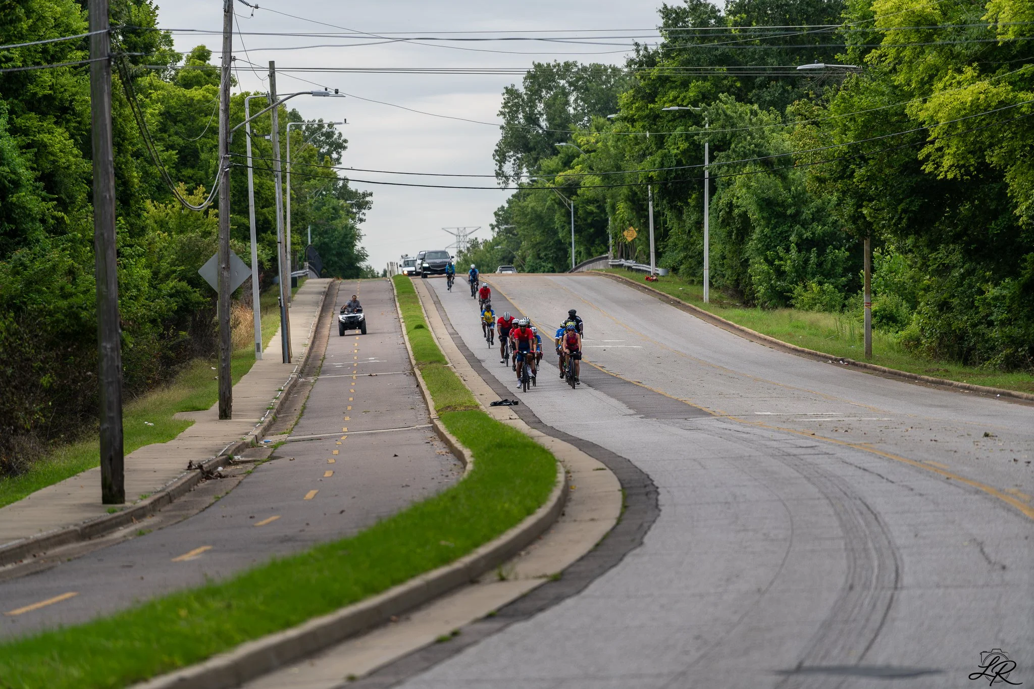 A group of cyclists riding on a divided road with cars and a scooter following behind, surrounded by trees on a cloudy day.