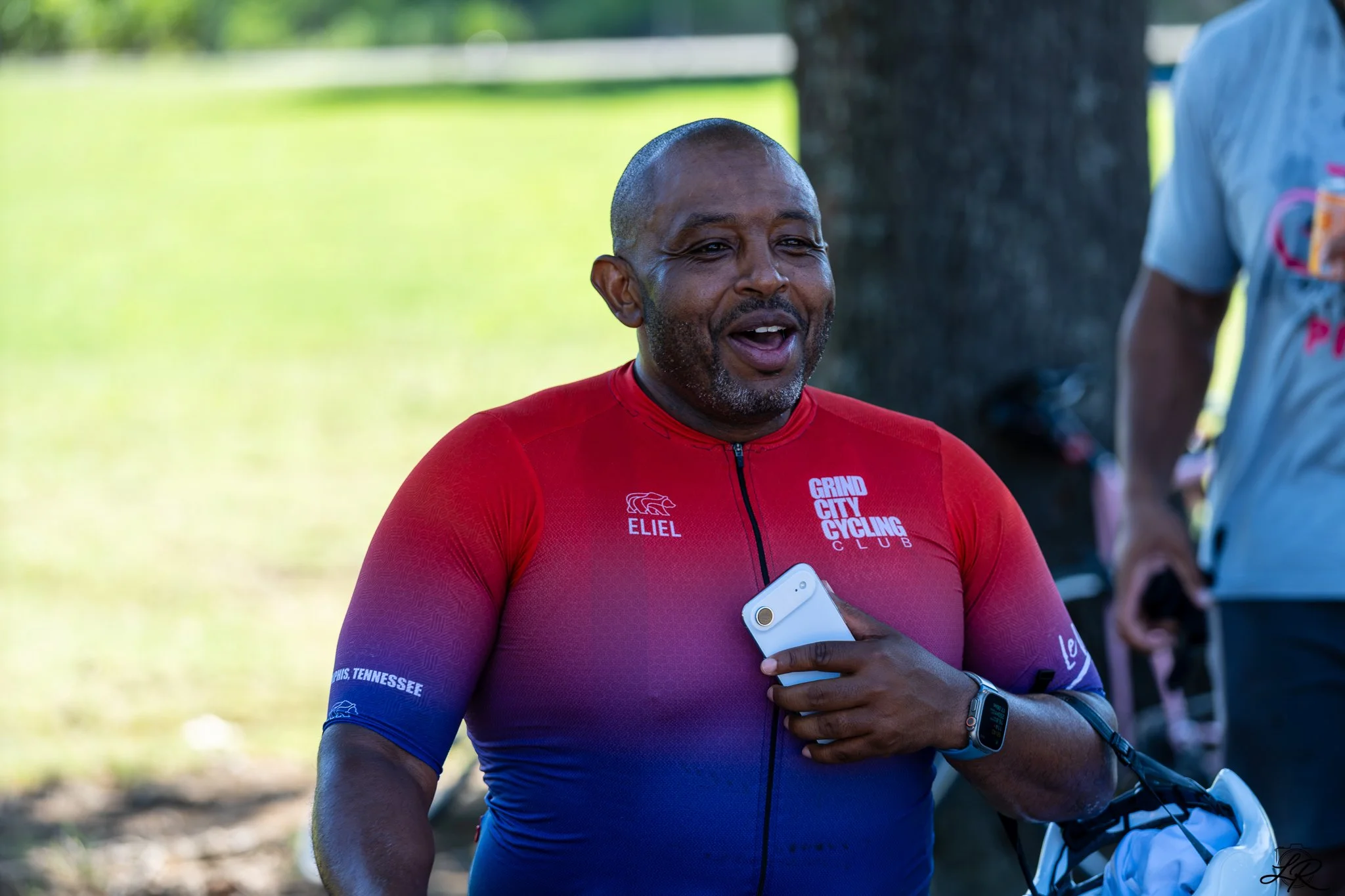 A man wearing a red and blue cycling jersey is smiling outdoors while holding a smartphone. He is at a cycling event, with a blurred person and trees in the background.