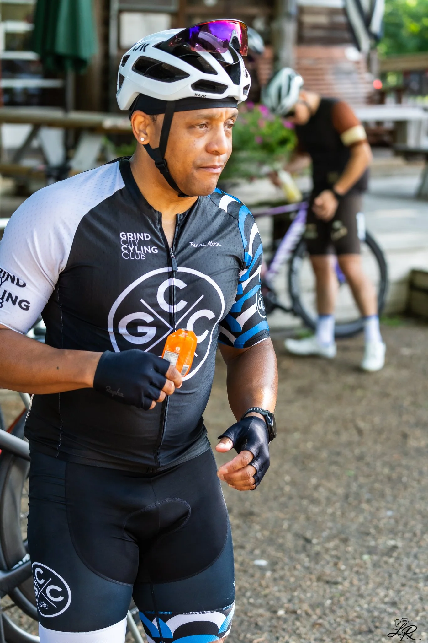 A man in cycling gear, wearing a white cycling helmet with magnified lenses, and a black cycling jersey with blue, white, and black accents, standing outdoors. In the background, another cyclist in black gear is adjusting their bicycle near wooden st