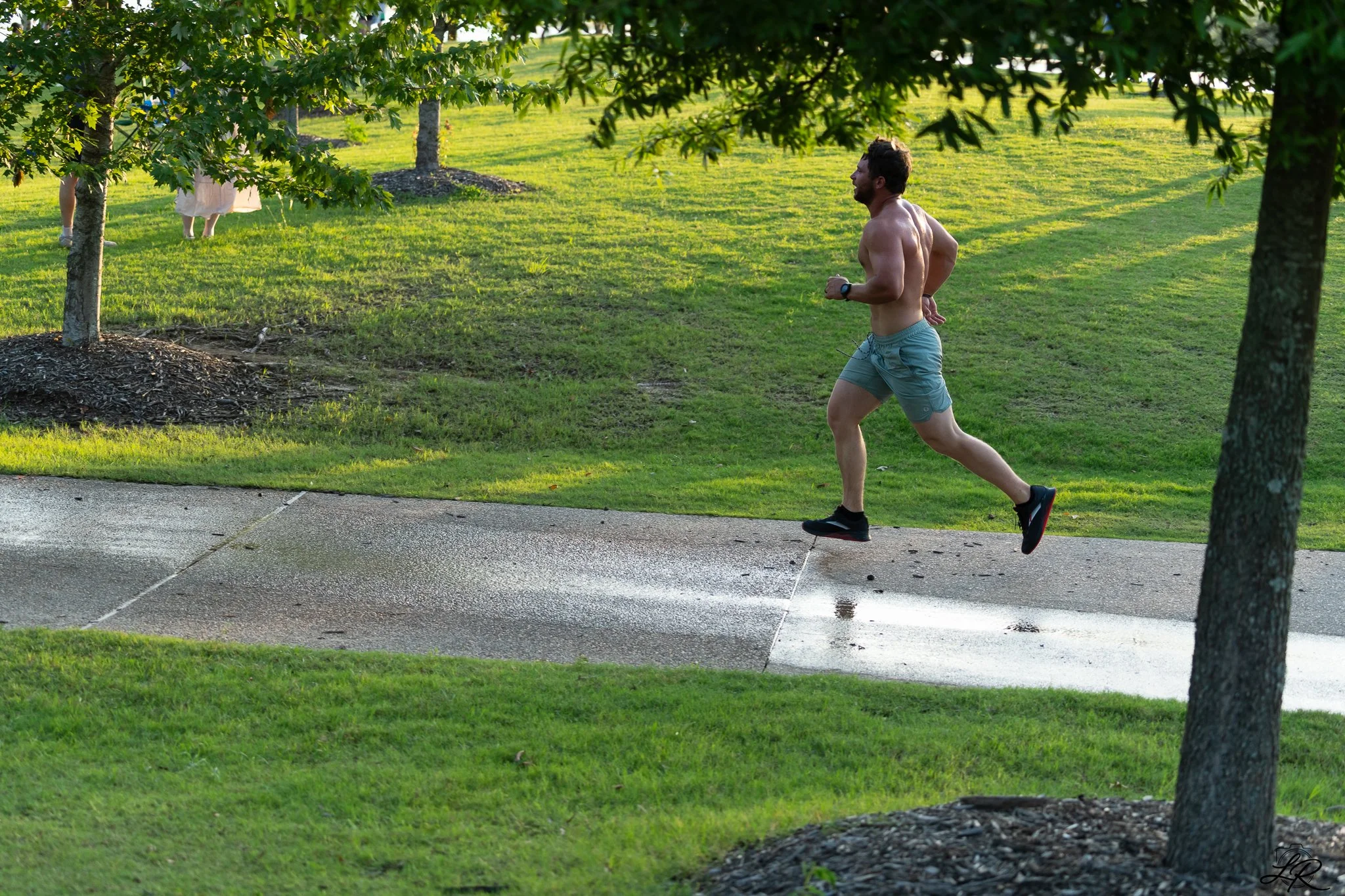 A man jogging shirtless on a park pathway, with trees and grass in the background, and two children partially obscured by tree leaves.