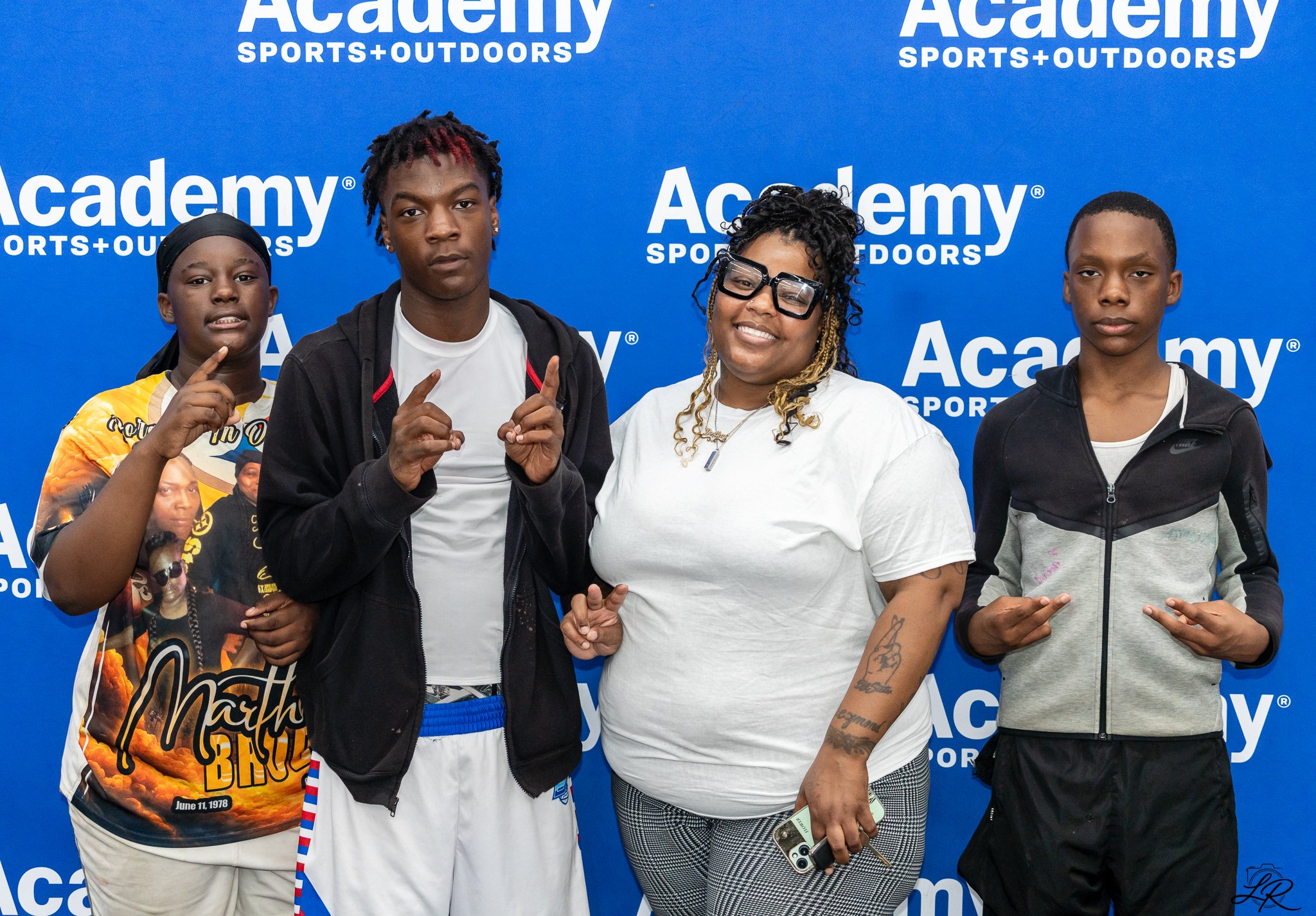 Four people standing in front of a blue backdrop with white text that reads "Academy Sports+Outdoors." They are posing for the photo, some making hand gestures. The group includes two young men, a young woman, and an older woman smiling.