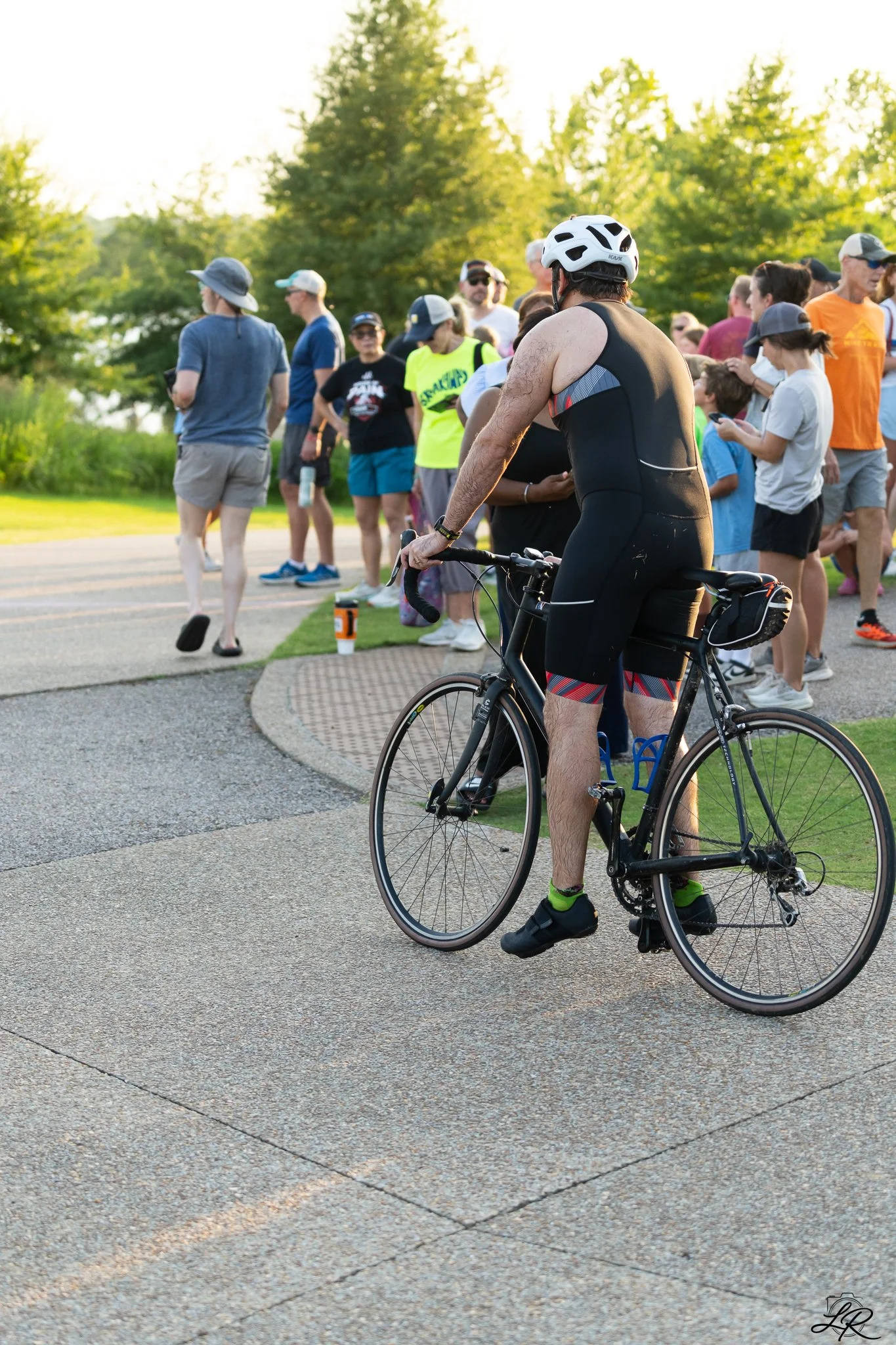 A man in a black cycling outfit and a white helmet is holding a black road bicycle, while a group of people, including children and adults, gather in the background outdoors during the daytime, with trees and greenery around.