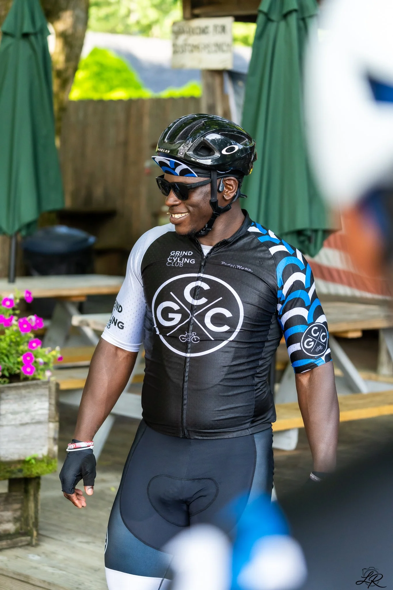 A man wearing a black bicycle helmet, black sunglasses, and cycling gear, smiling as he rides his bike.