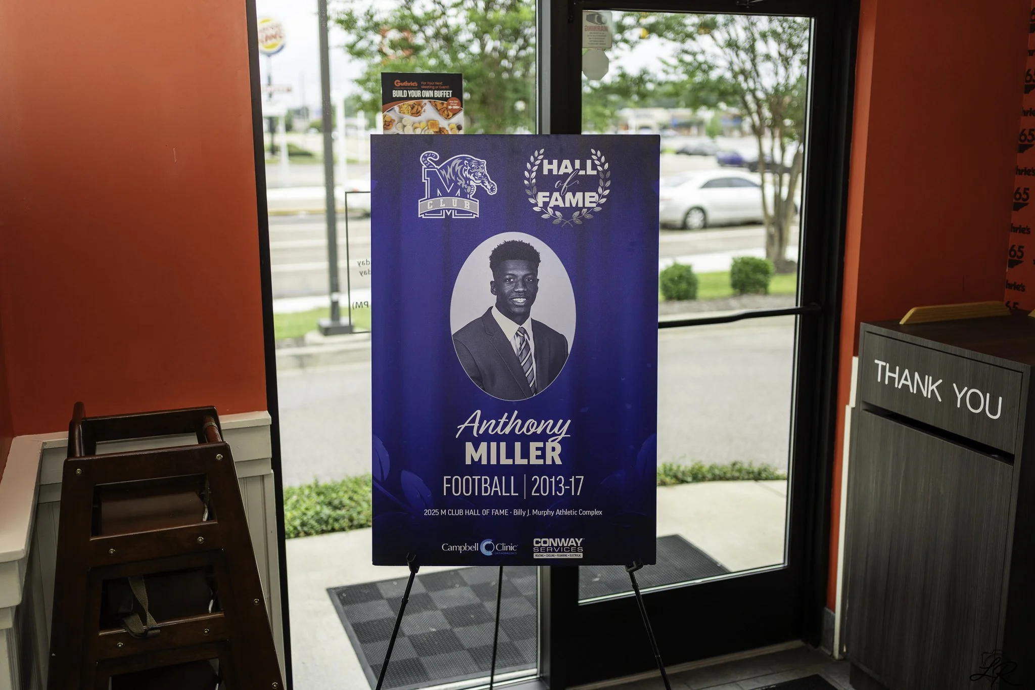 A poster inside a building features a young man in a suit and tie, with text indicating he is Anthony Miller, a football player from 2013 to 2017. The poster is part of the Hall of Fame, sponsored by Campbell Clinic and Conway Services, and is locate