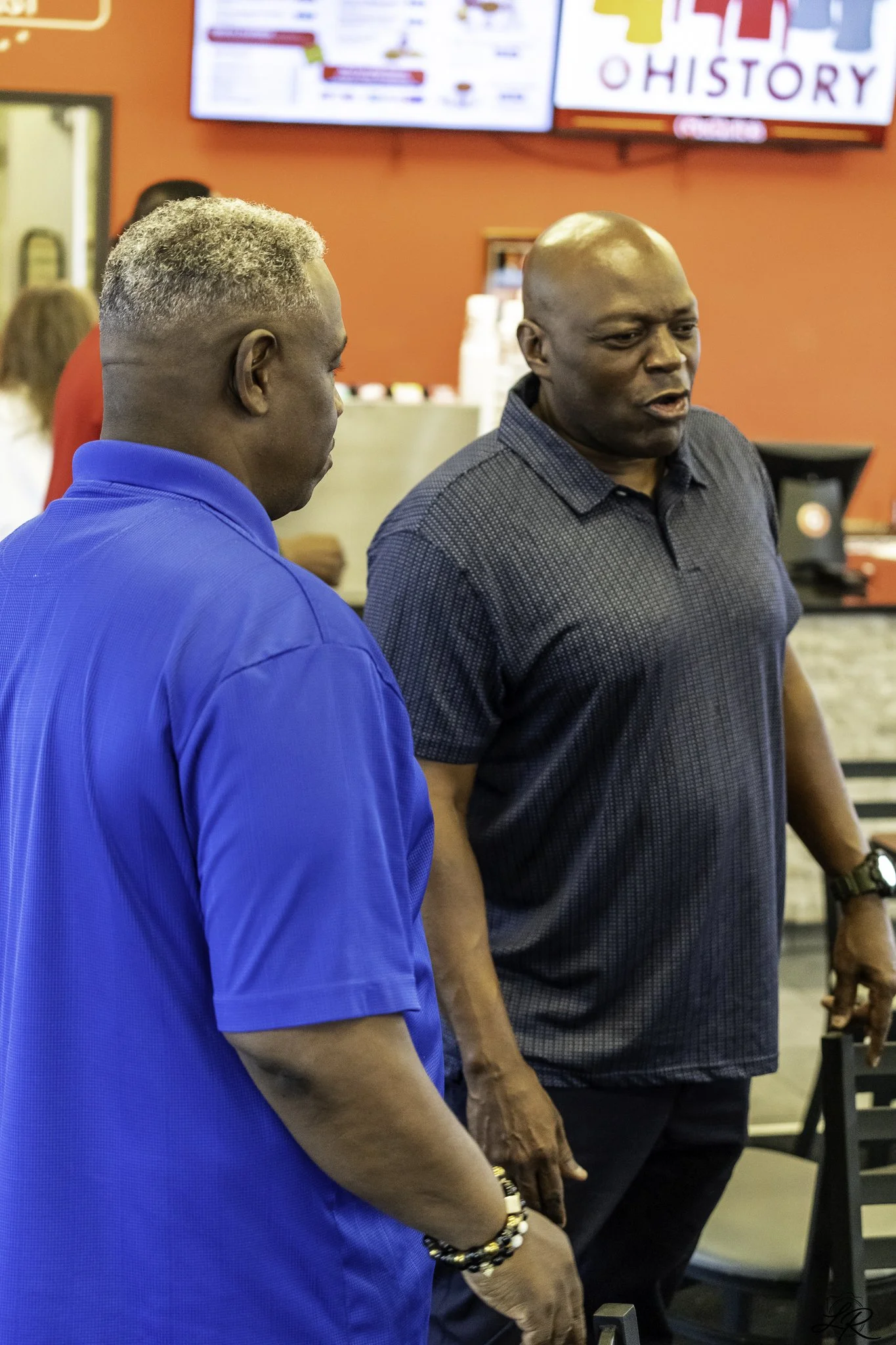 Two men are standing indoors, engaged in conversation. One man is wearing a bright blue shirt and has a short, curly hairstyle with gray and black hair. The other man is bald, wearing a dark gray, short-sleeved, button-up shirt. They are in front of 
