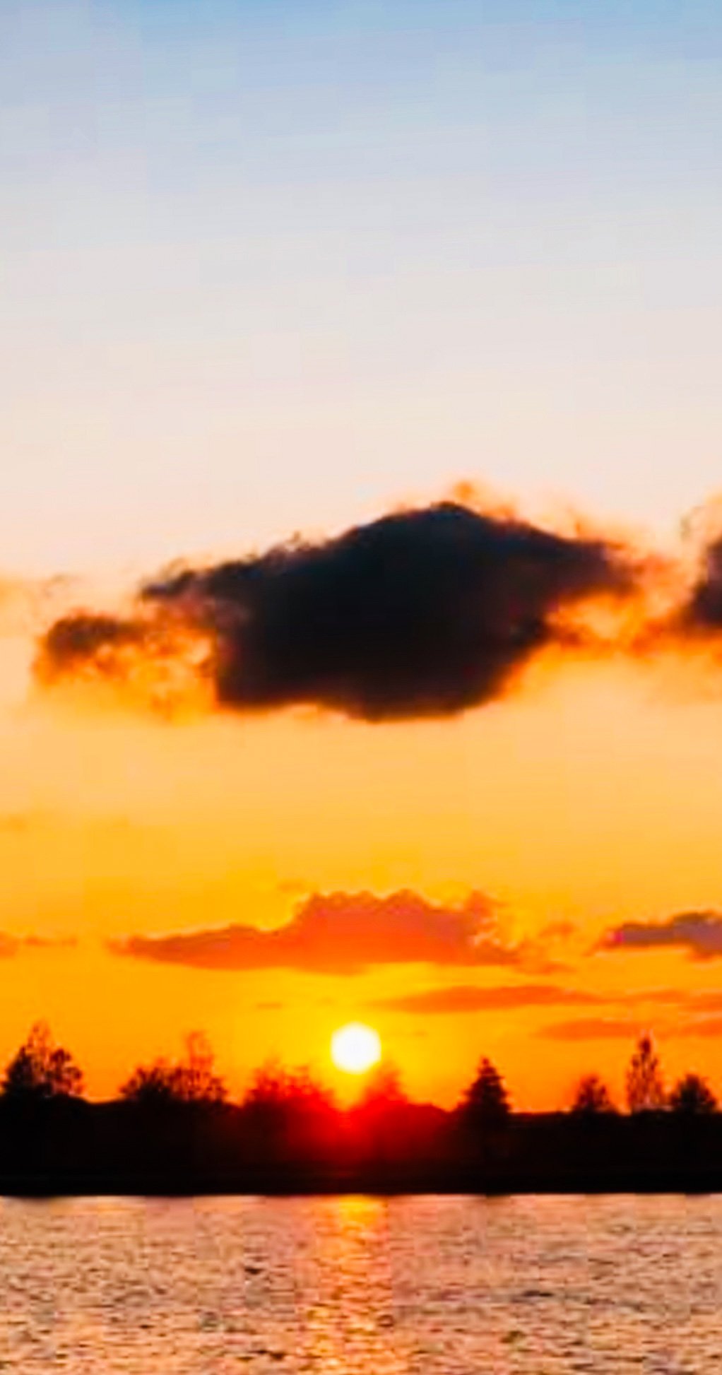 Sunset over a body of water with orange and yellow sky, clouds, and silhouettes of trees in the distance.