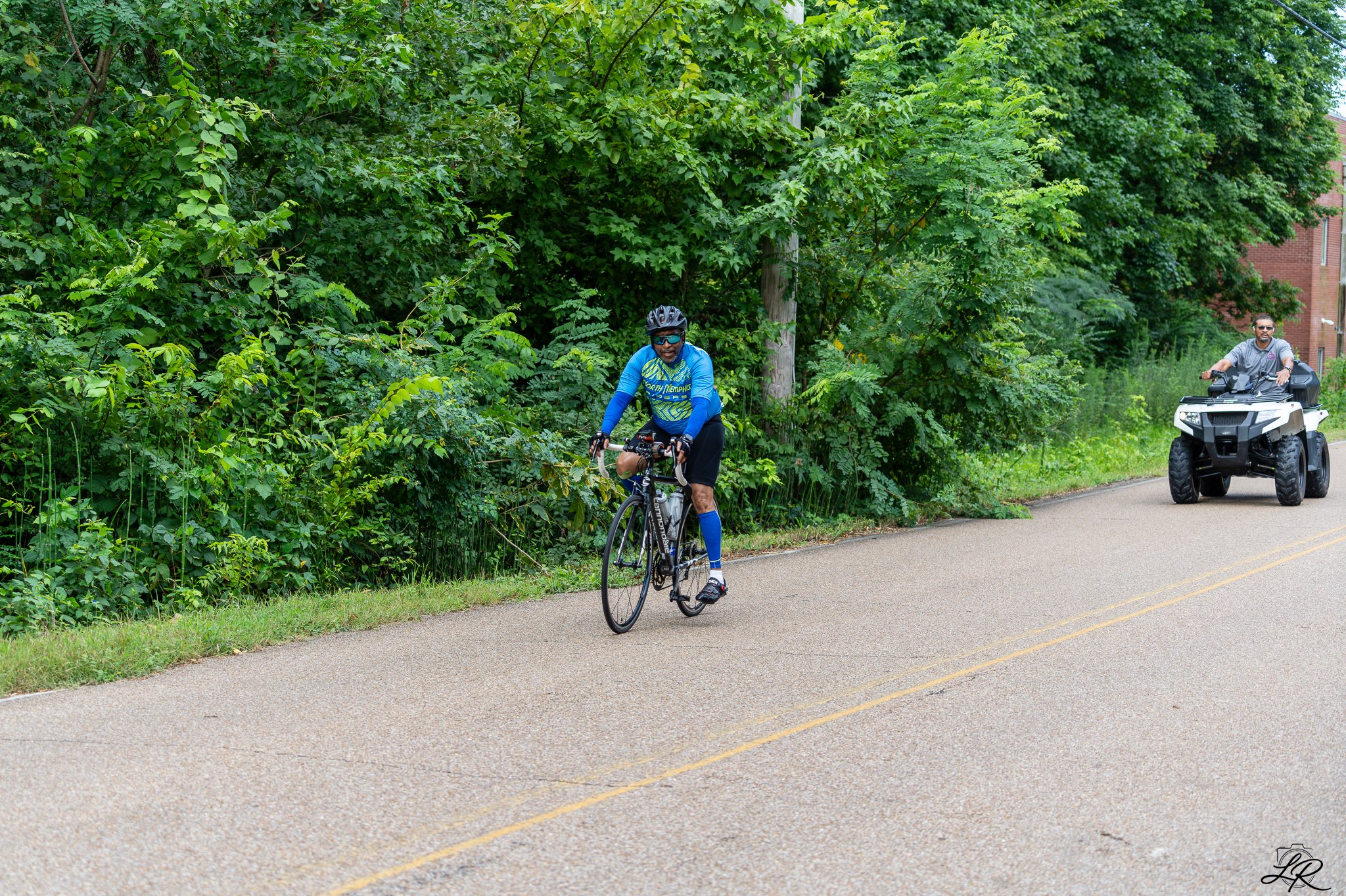 A man riding a bicycle along a paved road next to dense green trees, with another man riding a motorized quad bike in the background.