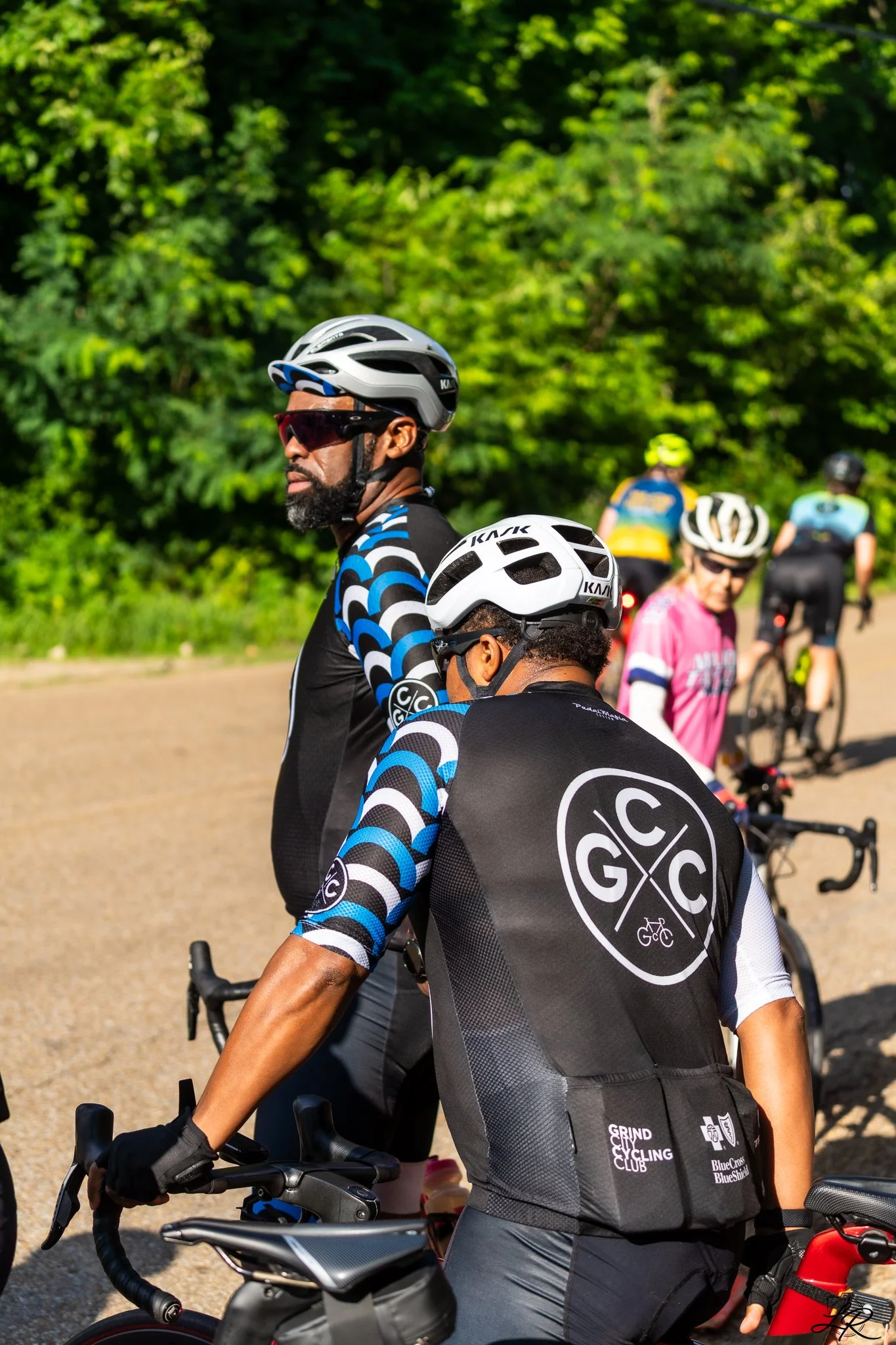 Group of cyclists, with focus on a man in the foreground wearing a black and blue cycling jersey, a gray helmet, sunglasses, and riding a black bicycle, with green trees in the background.