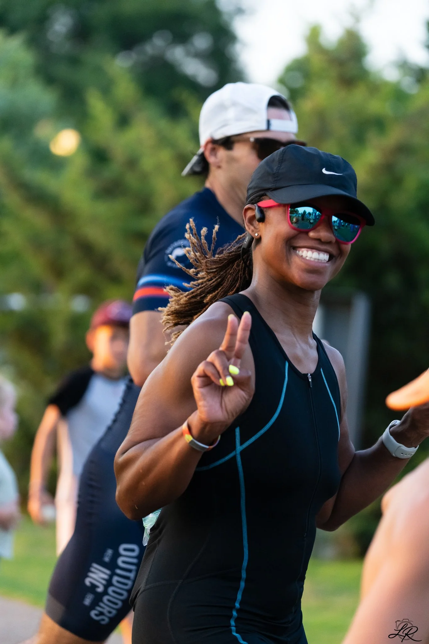 Smiling woman in black athletic wear and sunglasses running in a race, with people in the background.