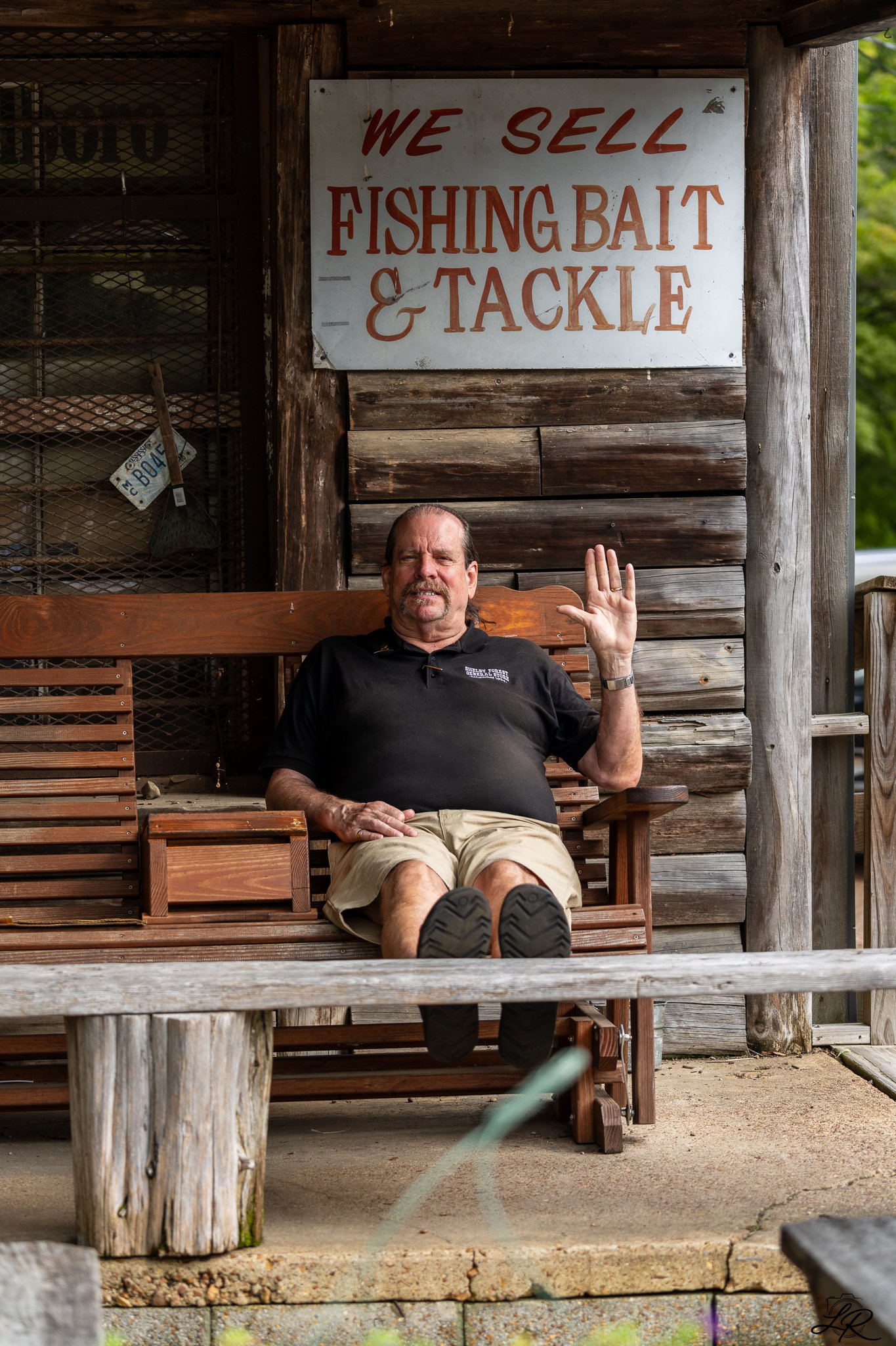 A man sitting on a wooden bench under a cabin, waving with one hand, with a sign behind him that reads, "We sell fishing bait & tackle."