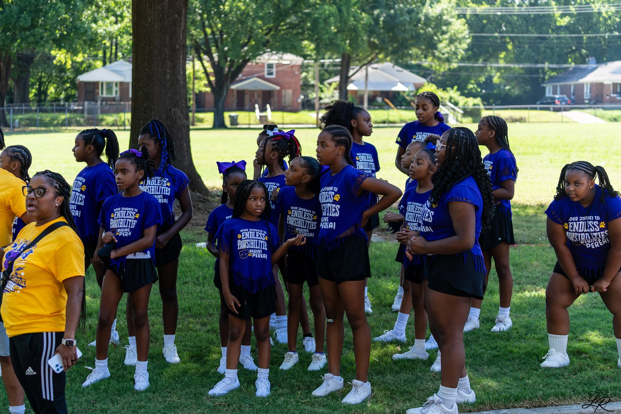 Group of young girls in athletic uniforms standing outdoors in a park, with trees and residential houses in the background.