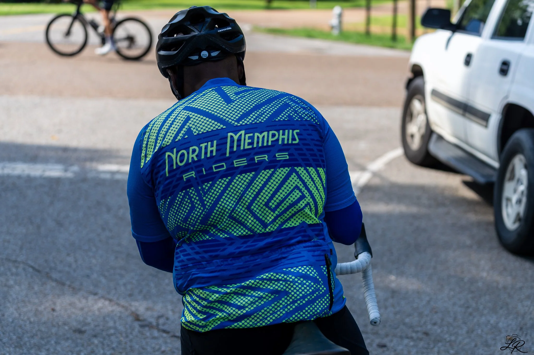 A person wearing a blue and green cycling jersey with the words "North Memphis Riders" on the back, a black helmet, and black pants, holding a white bicycle handlebar, with a blurred cyclist in the background, near a parked white vehicle in an outdoo