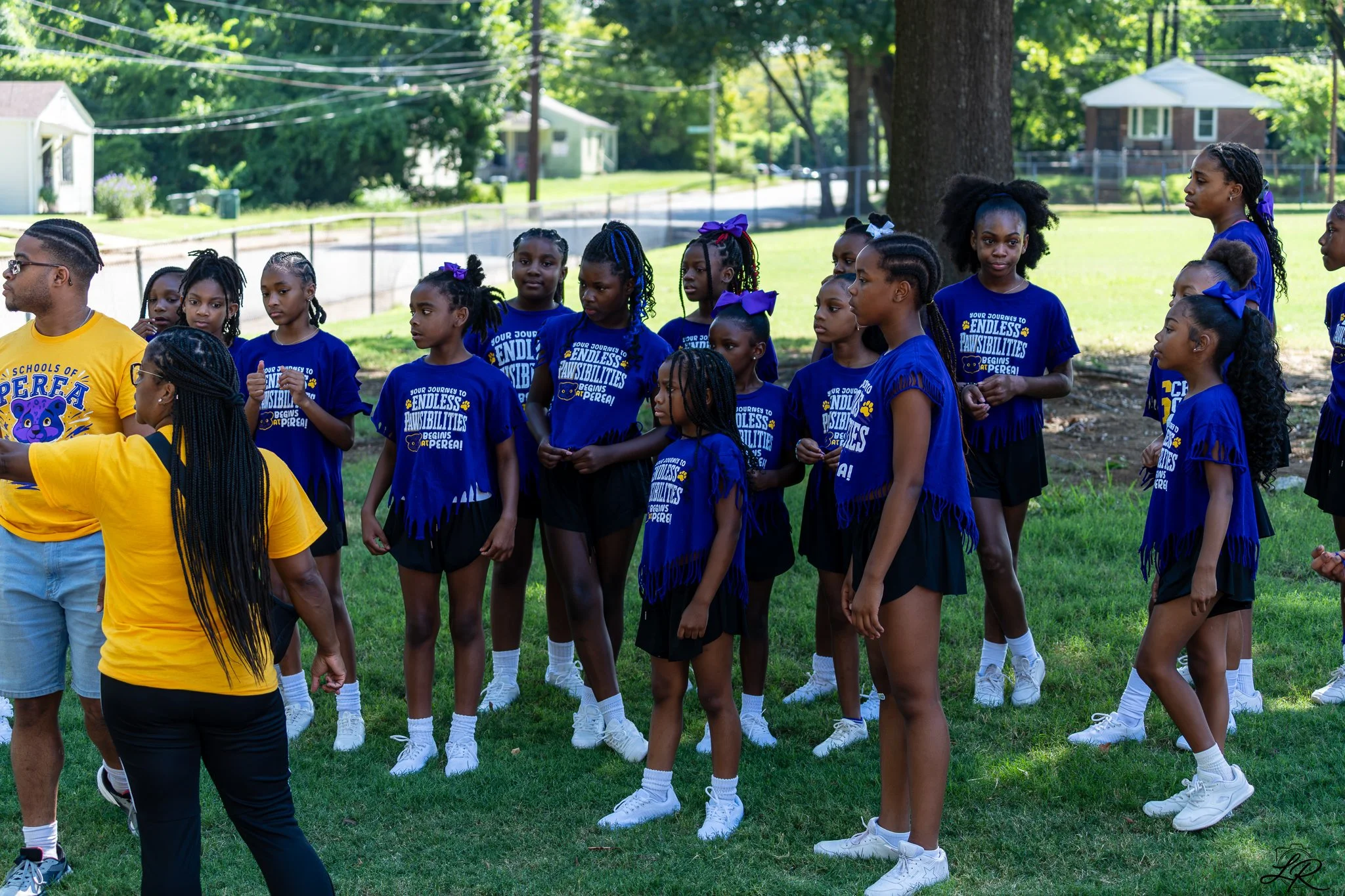 A group of girls dressed in matching blue t-shirts and black skirts, standing outdoors in a park-like setting with trees and houses in the background. Some girls have bows in their hair, and their shirts have a logo and text about endless possibiliti