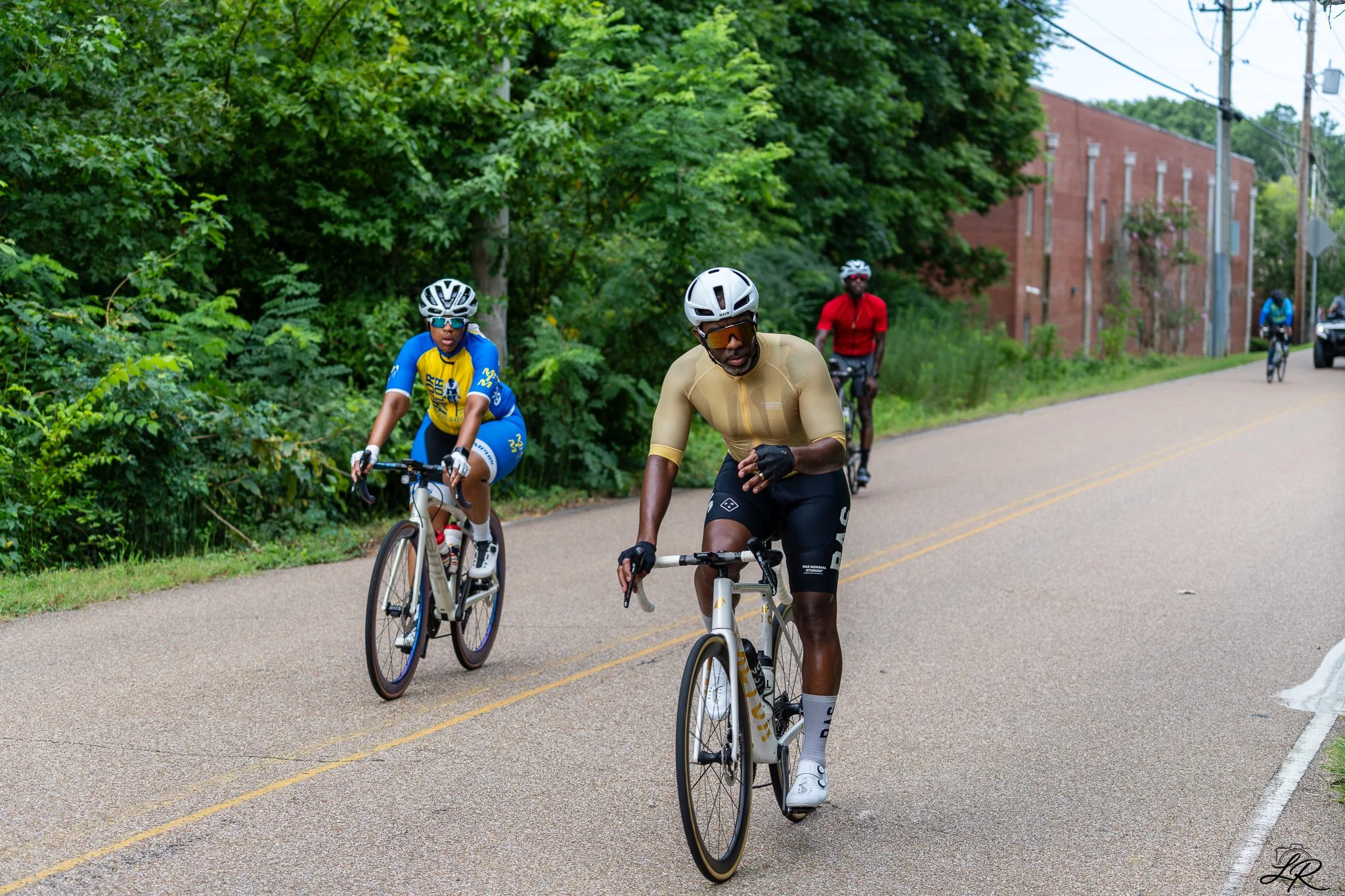 Four cyclists riding their bikes on a road with green trees and a red brick building in the background.