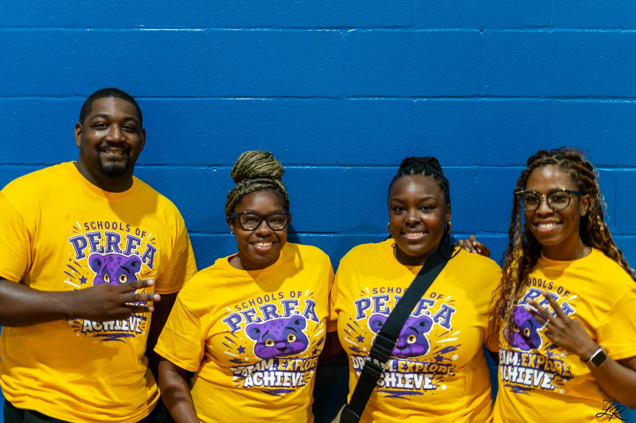 Group of five people wearing yellow t-shirts with a purple bear logo and the words 'TEAM EXPLORE ACHIEVE' standing against a blue wall, smiling at the camera.
