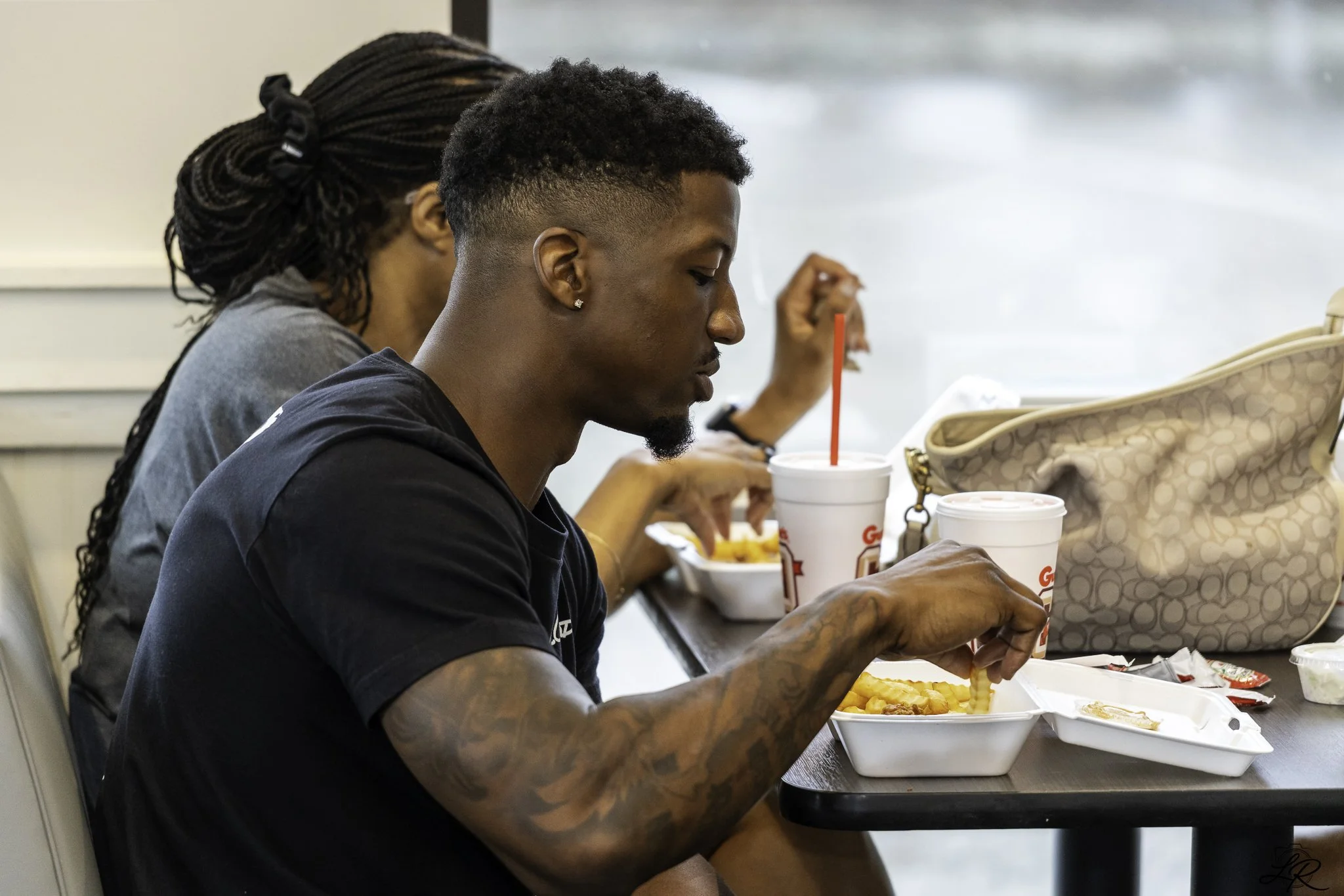 Two people are sitting at a table eating fast food. The person in the foreground has tattoos and is drinking from a large soda cup with a red straw. The person in the background has braided hair and is also eating.