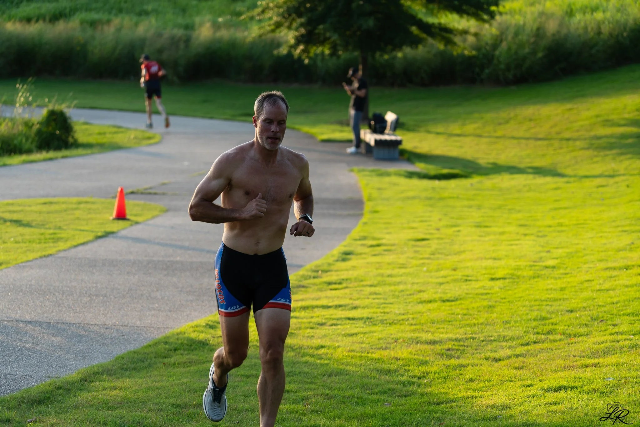 A shirtless man running outdoors on a paved path with green grass on each side and trees in the background, with a person in the distance and a person standing near a bench.
