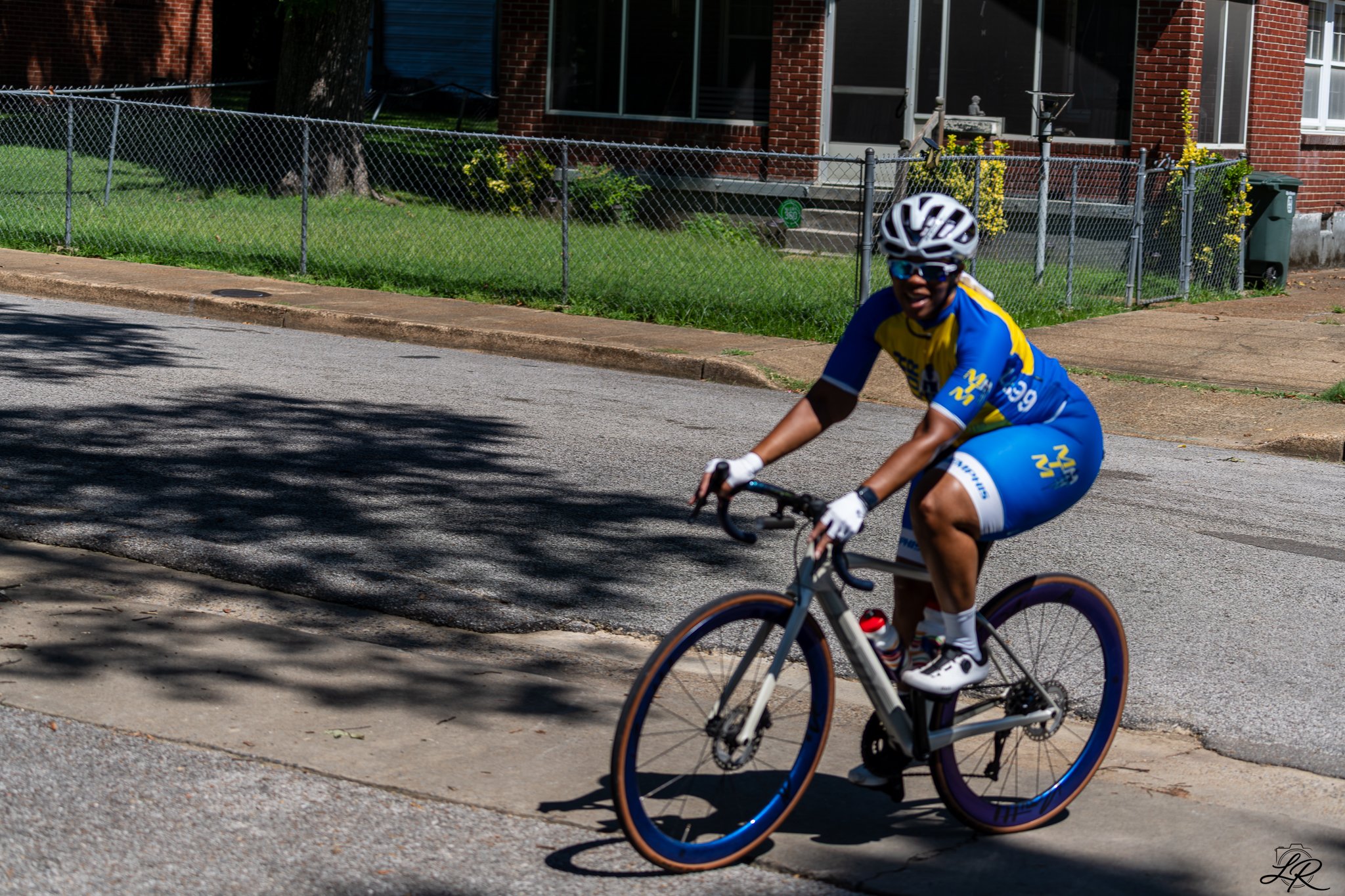 A cyclist wearing a helmet and sunglasses riding a bicycle on a suburban street, with a brick house, a green lawn, and a chain-link fence in the background.